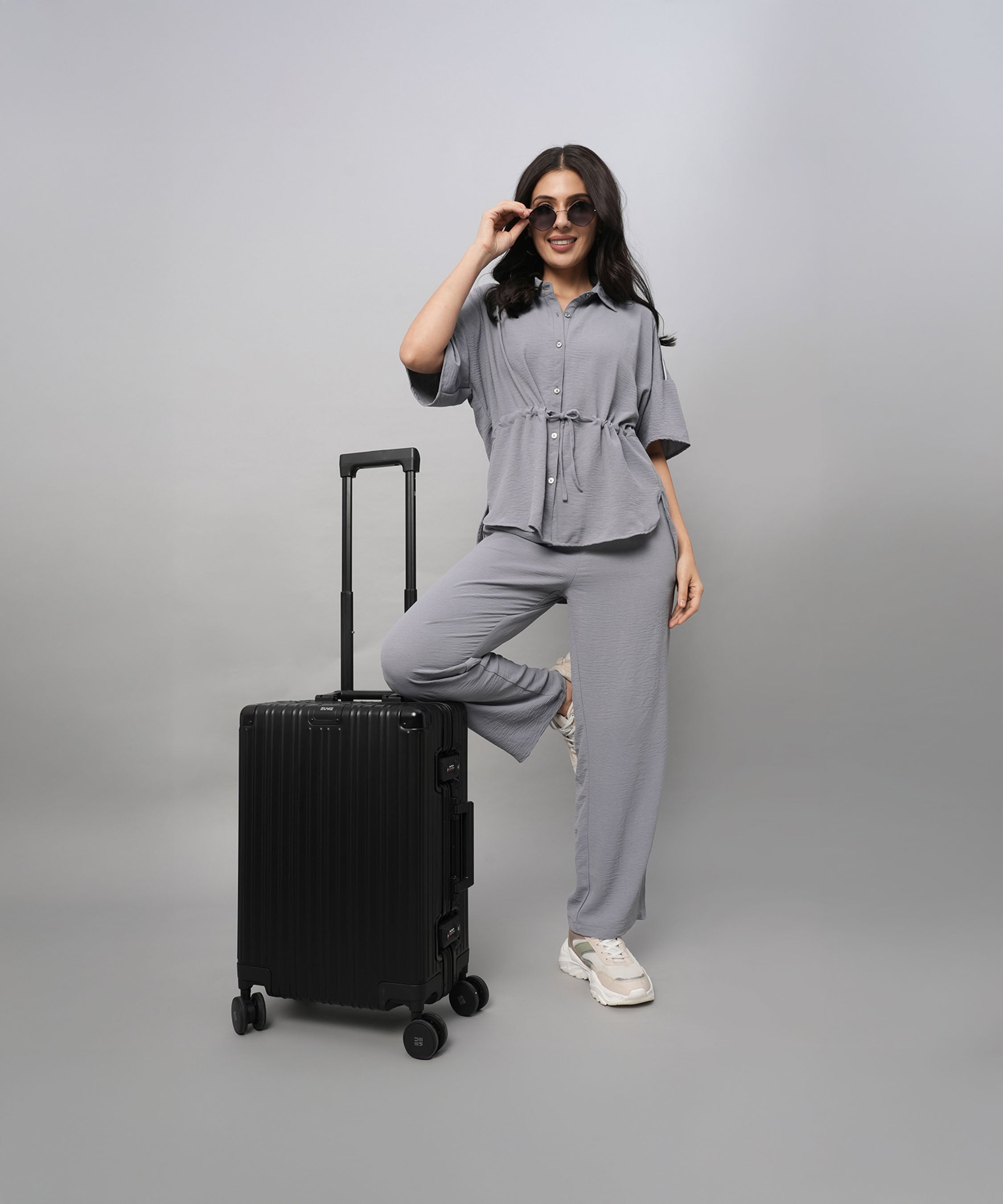 A woman poses confidently in a Grey Tie Up Top with Loose Pants Cord Set and sunglasses, one foot on a black suitcase, against a grey background.