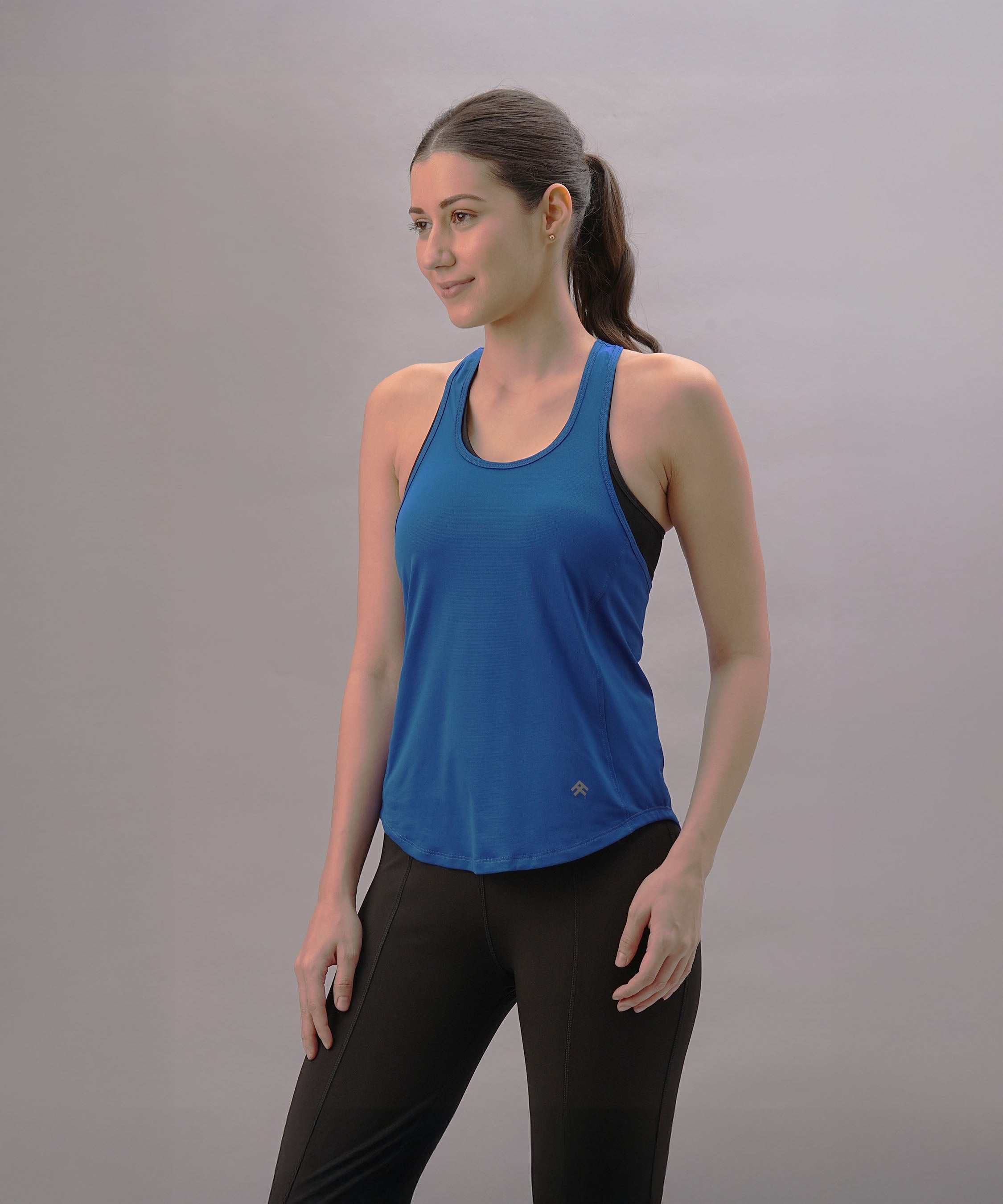 Woman wearing the Blue Racer Back Top and black pants stands against a plain light gray background, looking to the side.