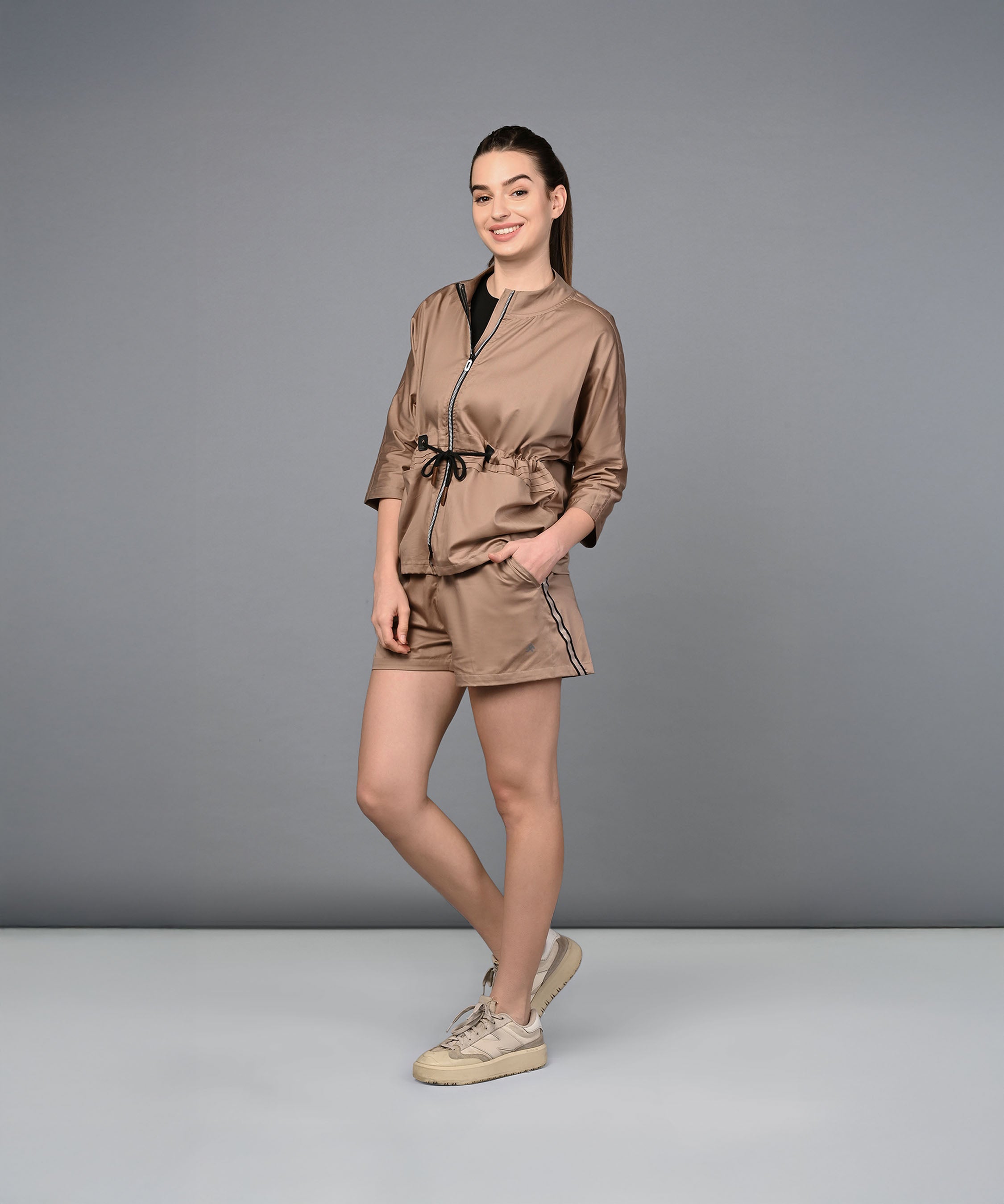 A woman stands smiling, wearing the Zipped Neutral Jacket Shorts Set with sneakers against a plain gray background.