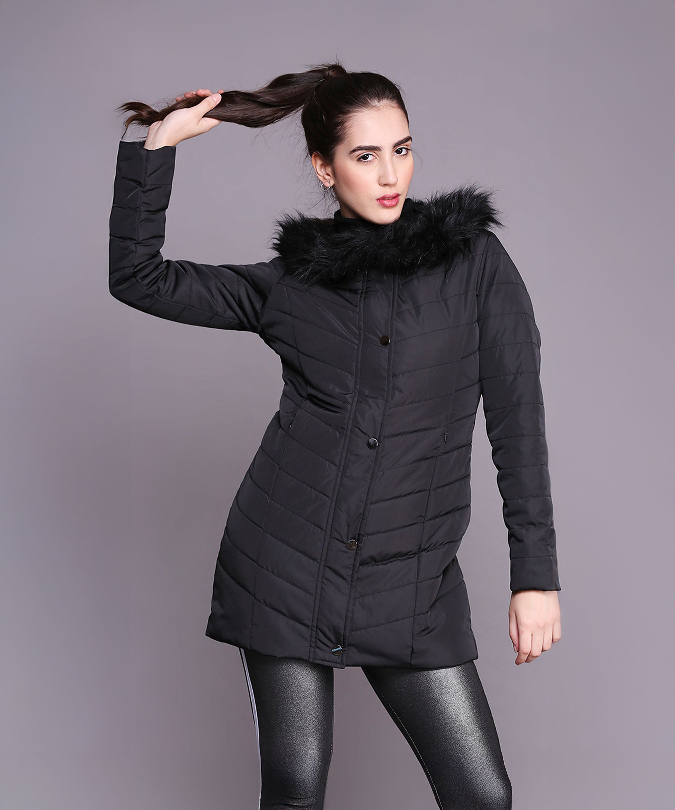 A woman strikes a pose against a plain background, holding her ponytail while wearing the Black Fur Collar Long Puffer Jacket With Button and Zip Details.