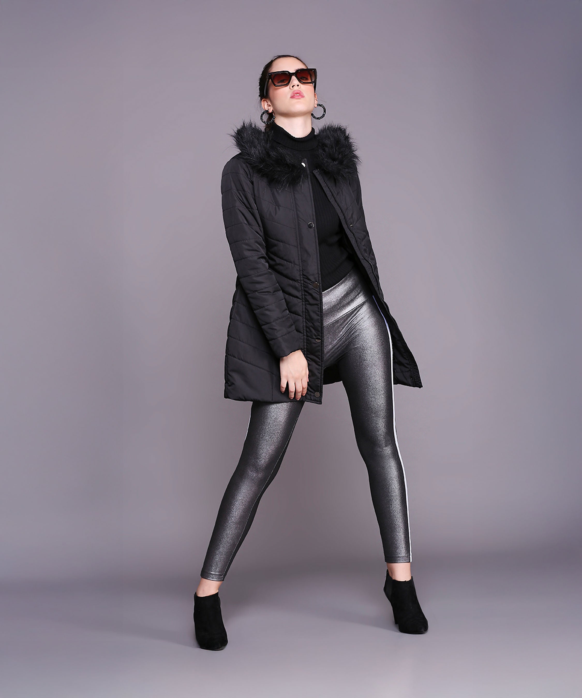 A woman confidently poses in a Black Fur Collar Long Puffer Jacket With Button and Zip Details, paired with metallic leggings, sunglasses, and ankle boots.