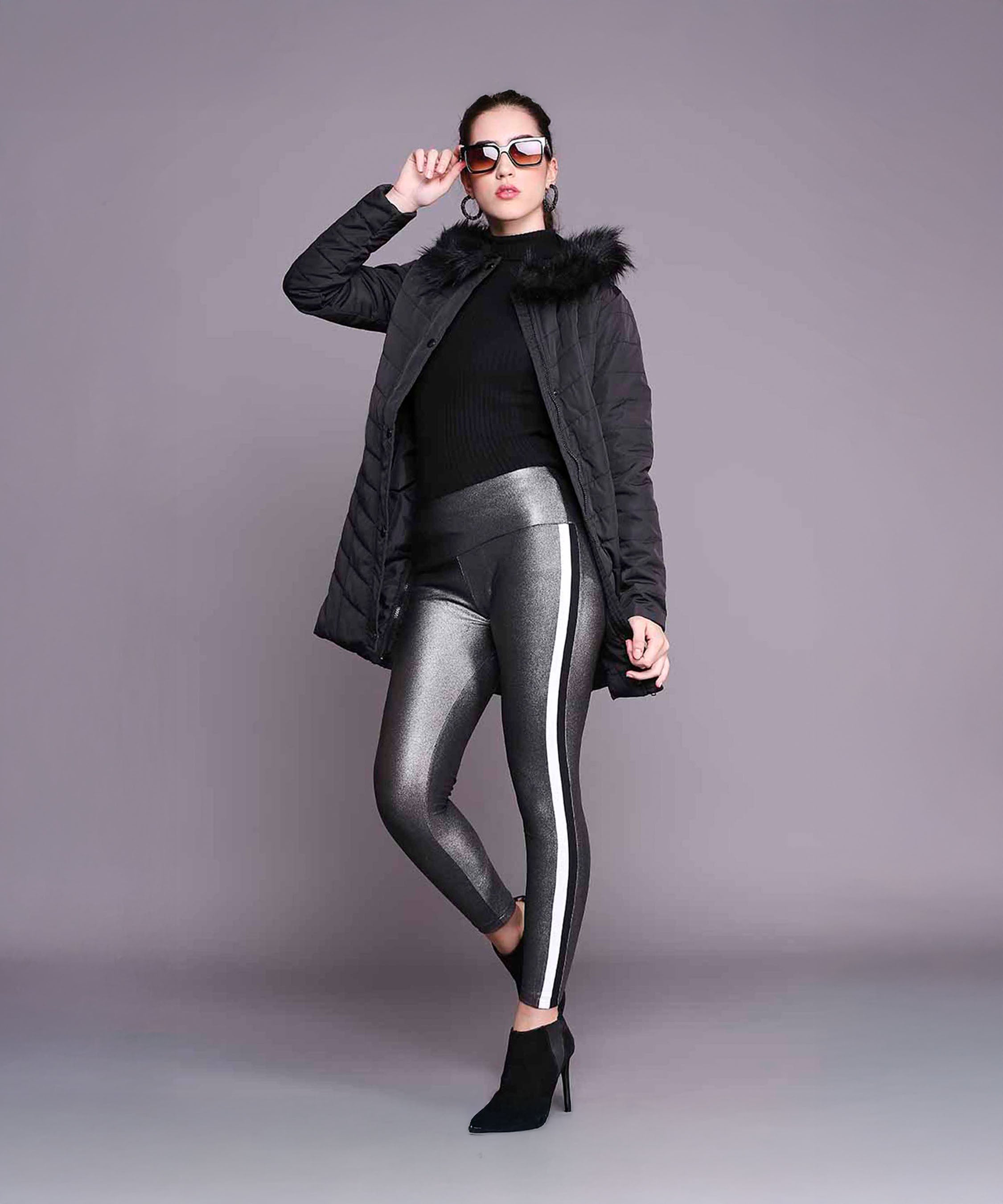 Woman in a Black Fur Collar Long Puffer Jacket With Button and Zip Details, metallic leggings, and sunglasses poses confidently against a plain gray background.