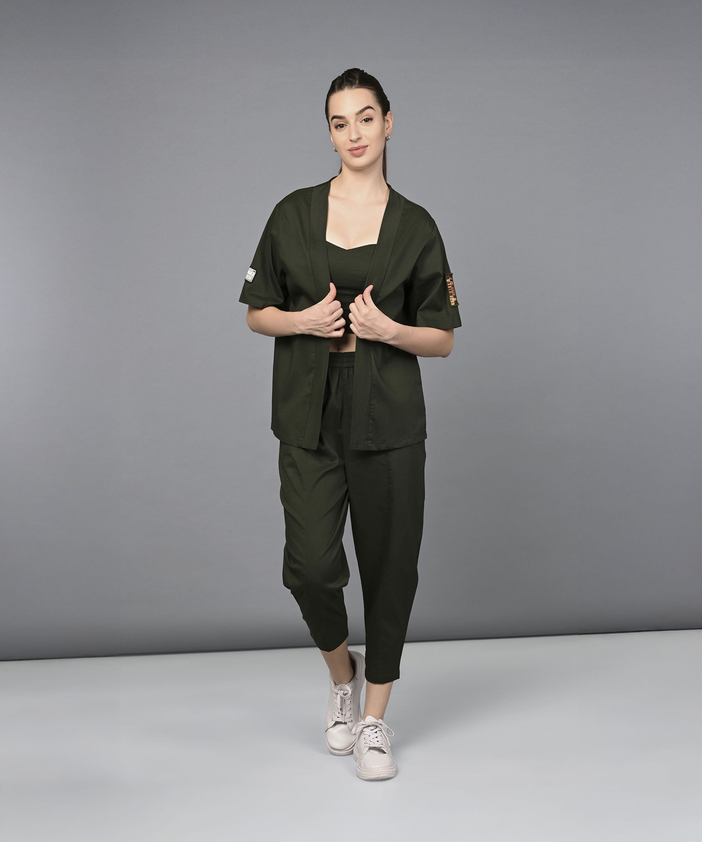 Woman in a Military Green Open Jacket & Yoga Pants with Bustiere set and white sneakers stands against a plain gray background.