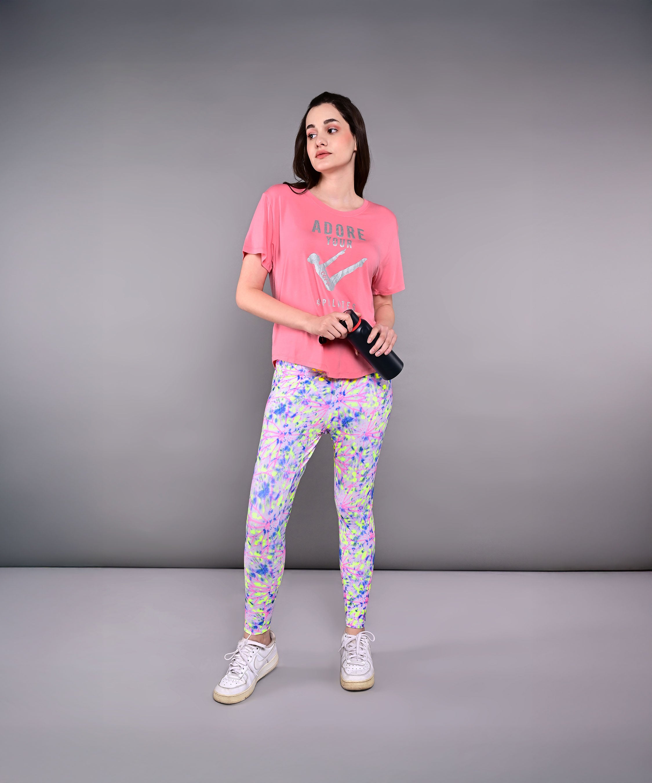 Woman in an 'Adore Your Core' printed tee and colorful leggings holds a water bottle, standing against a gray background.