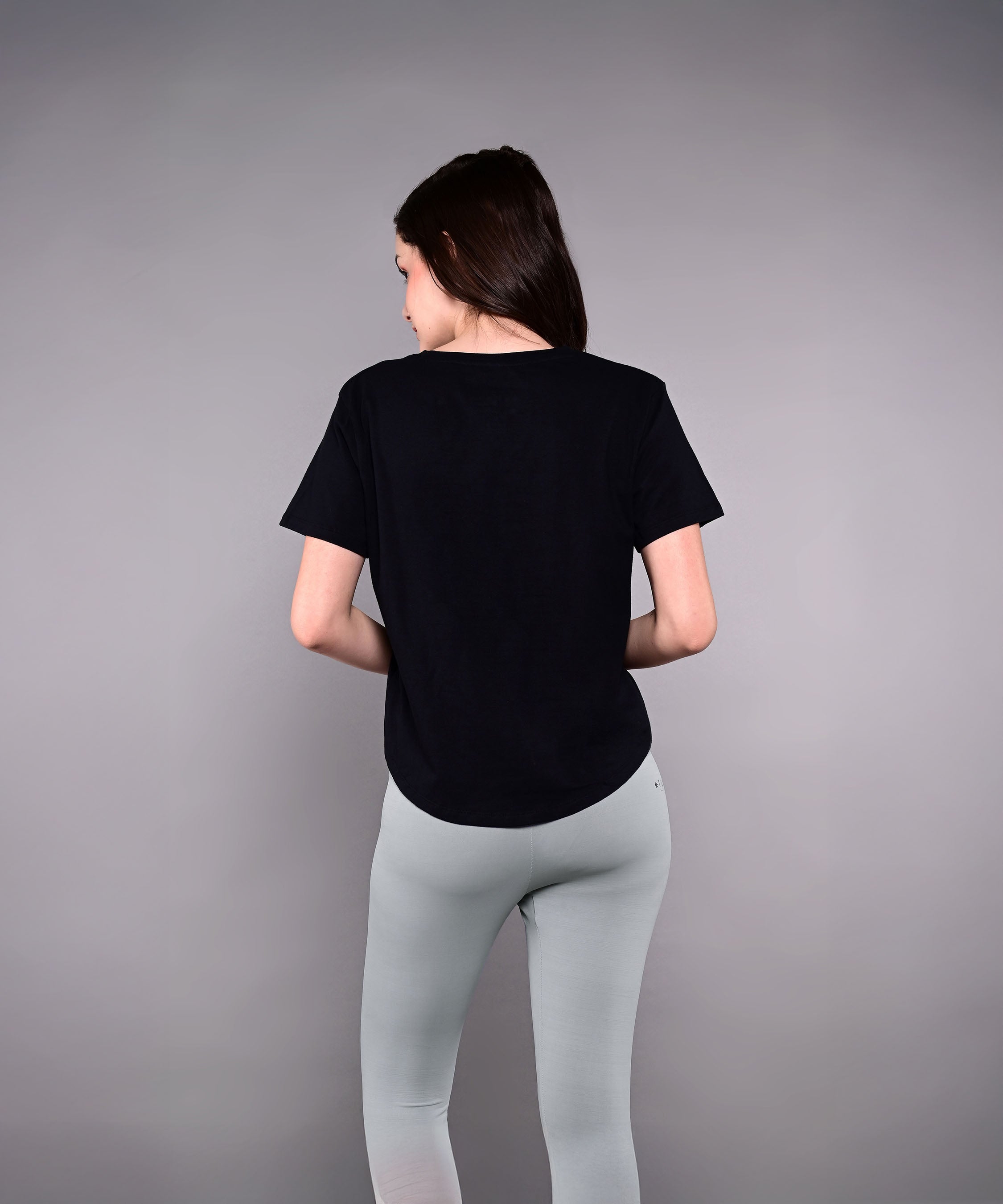 Woman with long dark hair stands with her back to the camera, wearing an 'Adore Your Core' Printed Tee and light gray leggings.