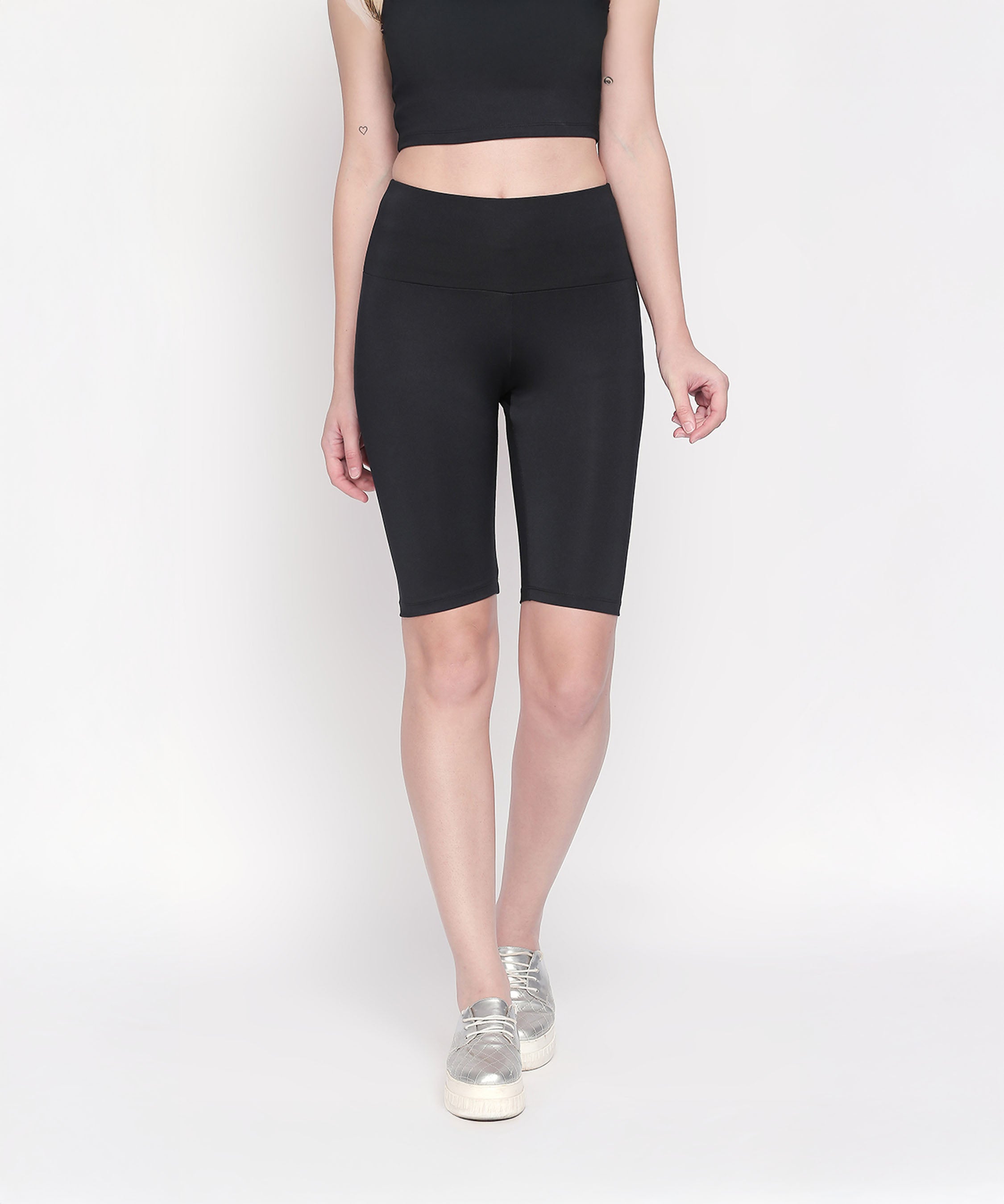 A woman stands against a white background wearing Black Biker/Cycling Shorts made from four-way stretch fabric, paired with a black crop top and silver sneakers.