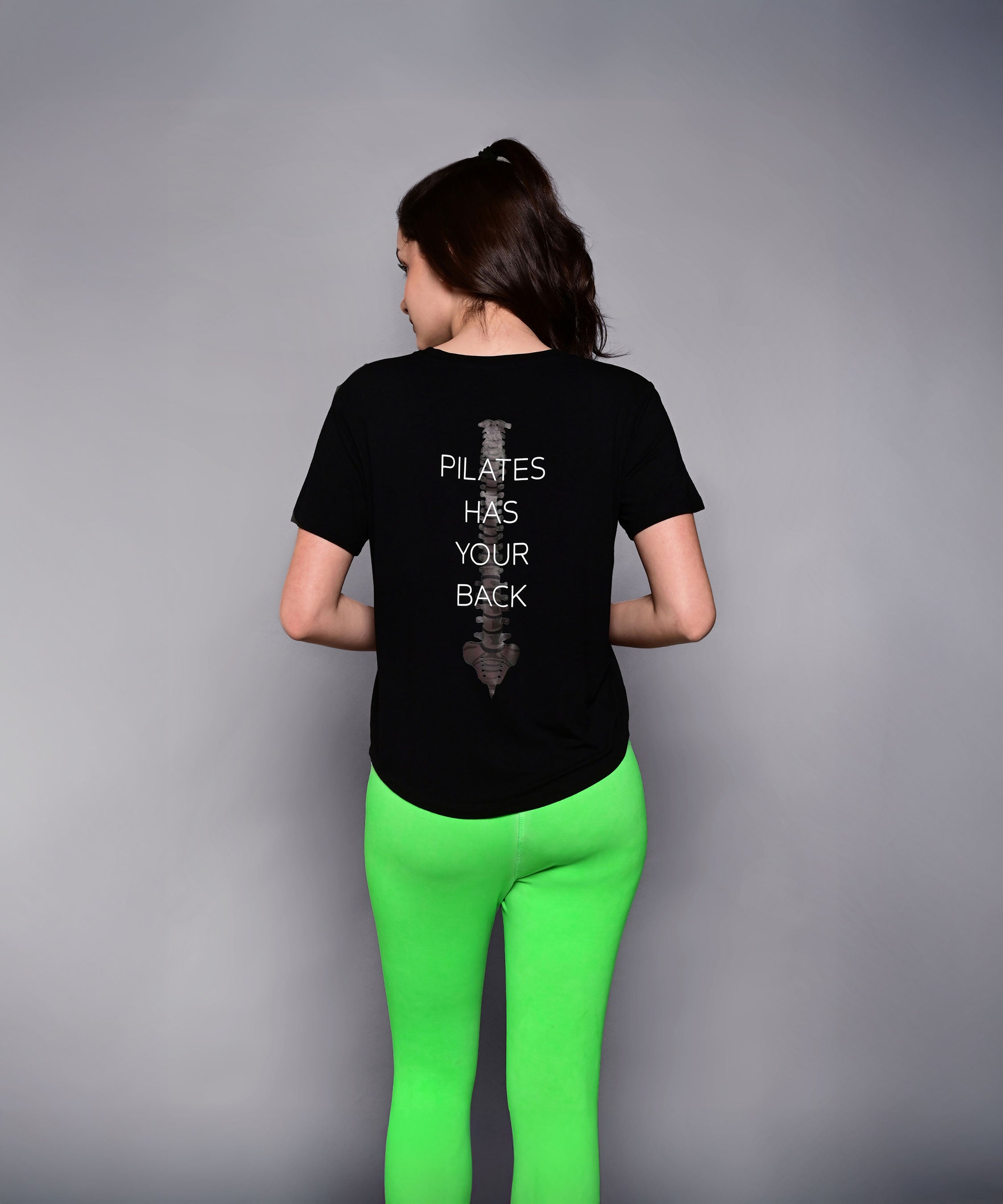 A woman in bright green leggings wears the 'Pilates Has Your Back' Printed Tee, featuring the motivational slogan on the back—perfect for workouts or casual wear.