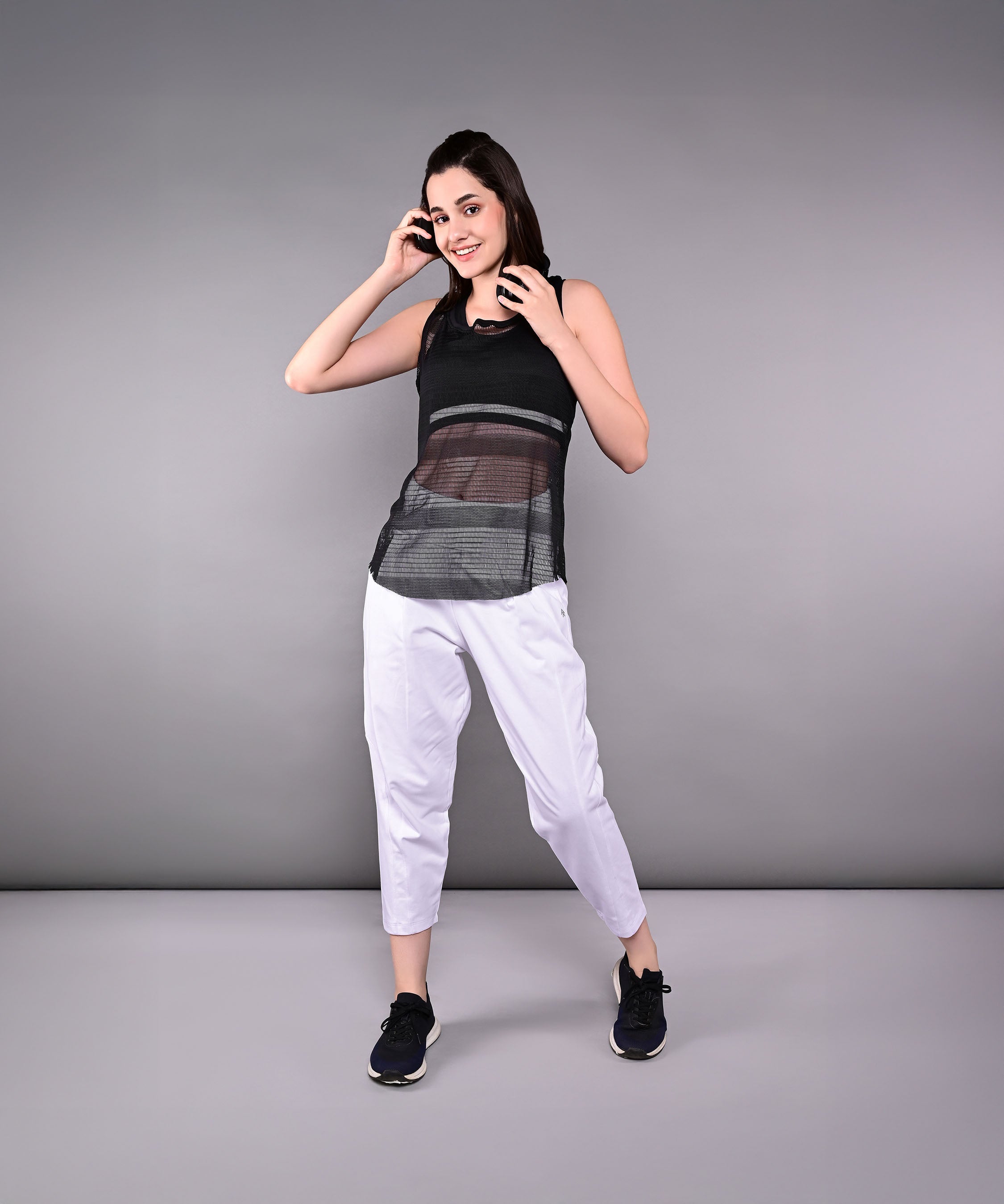 Woman smiles and poses in the Tuna Active Black Netted Tank Top with white pants against a plain gray background.