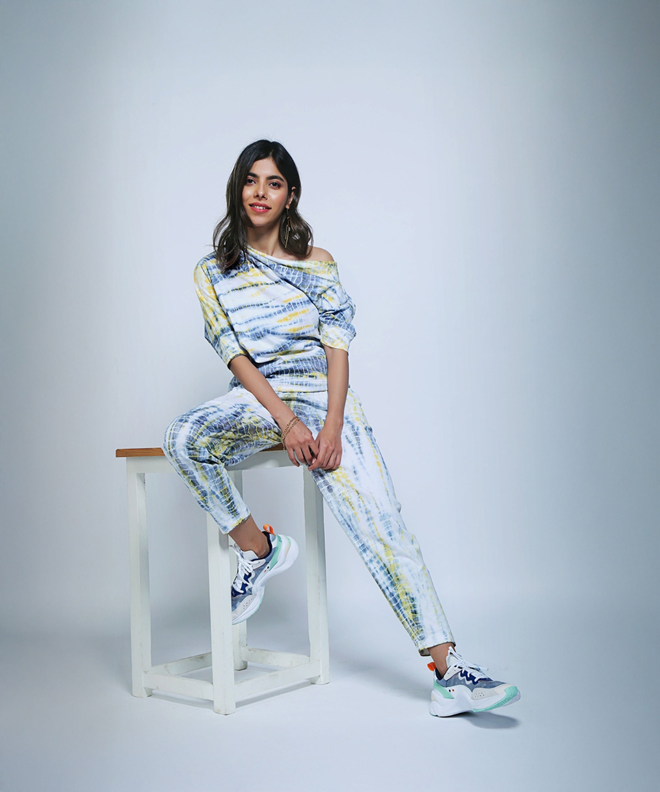 Woman in a yellow & grey One Side Off-Shoulder top and matching yoga pants sits smiling on a white stool in a studio.