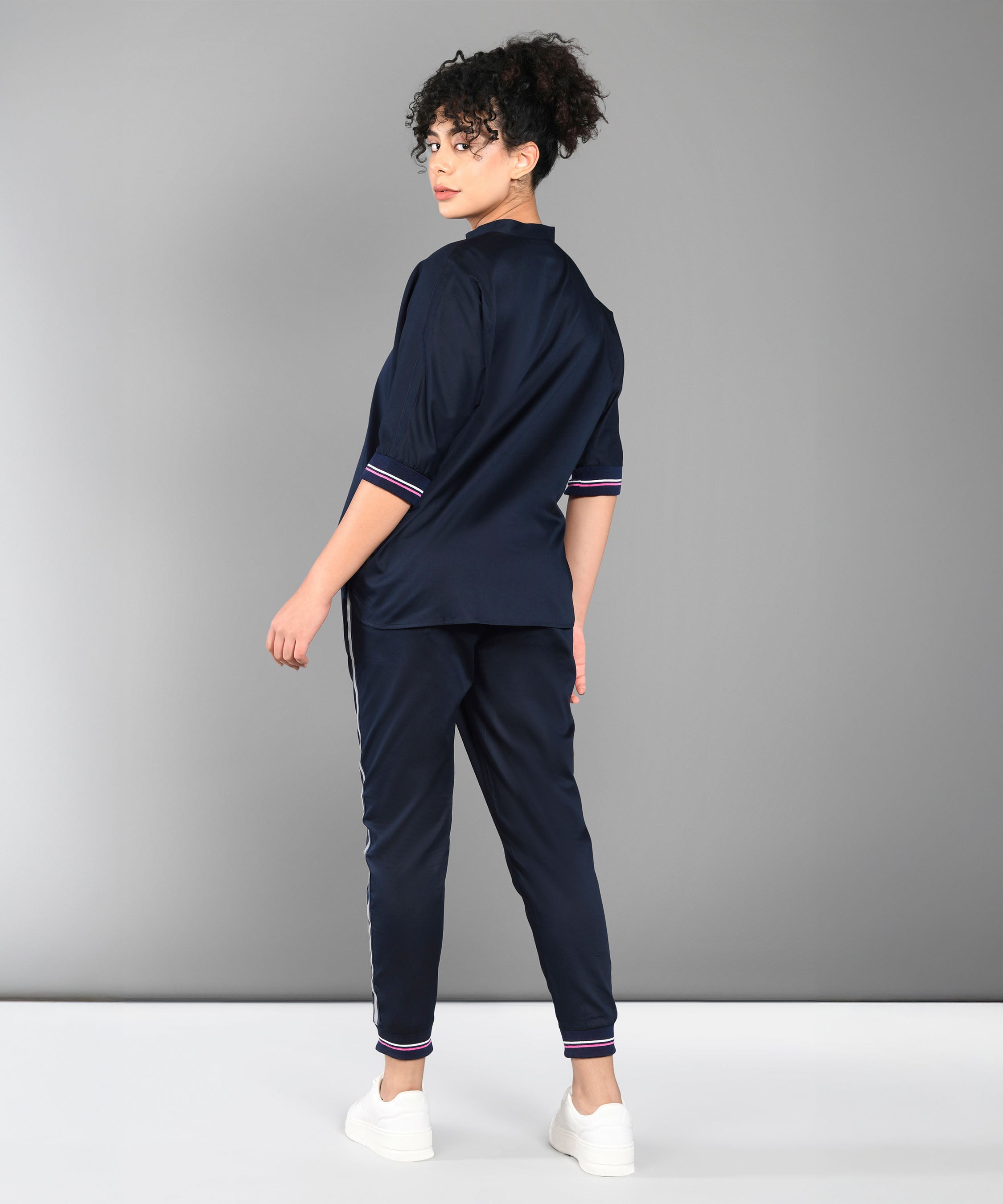 A woman in the Accented Set, a navy blue cotton co-ord, stands facing away and turns her head toward the camera, paired with white sneakers.