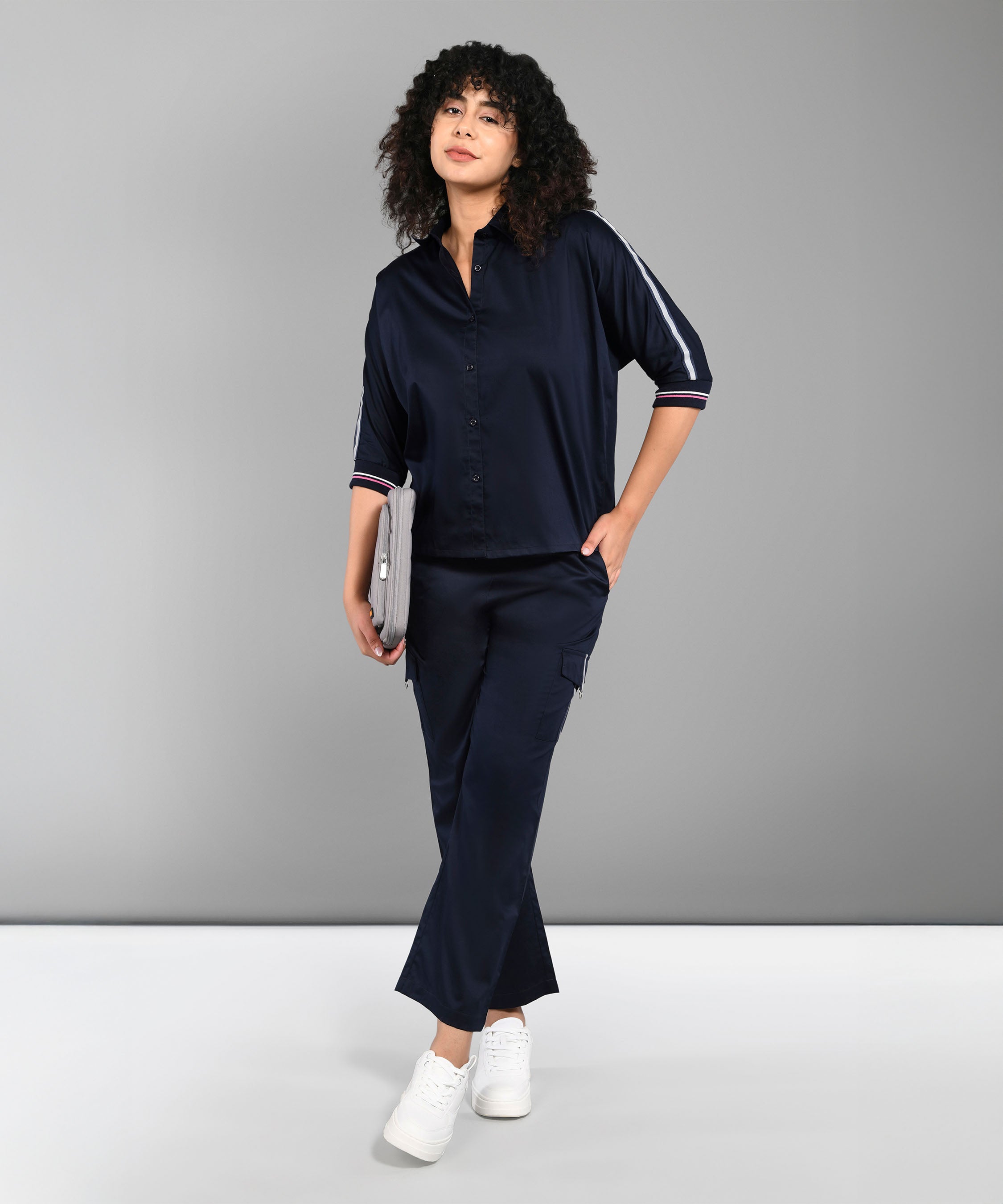 Woman with curly hair in the Navy Blue Chic Set and white sneakers, holding a tablet and standing against a gray background.