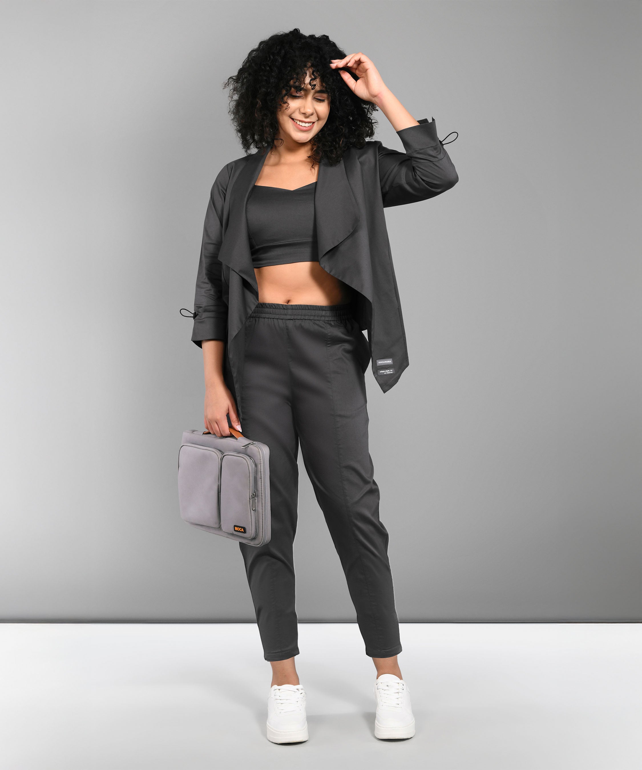 Woman in the Timeless Brilliance Grey Co-ord Set with Crop Top, holding a light gray bag and smiling against a gray background.
