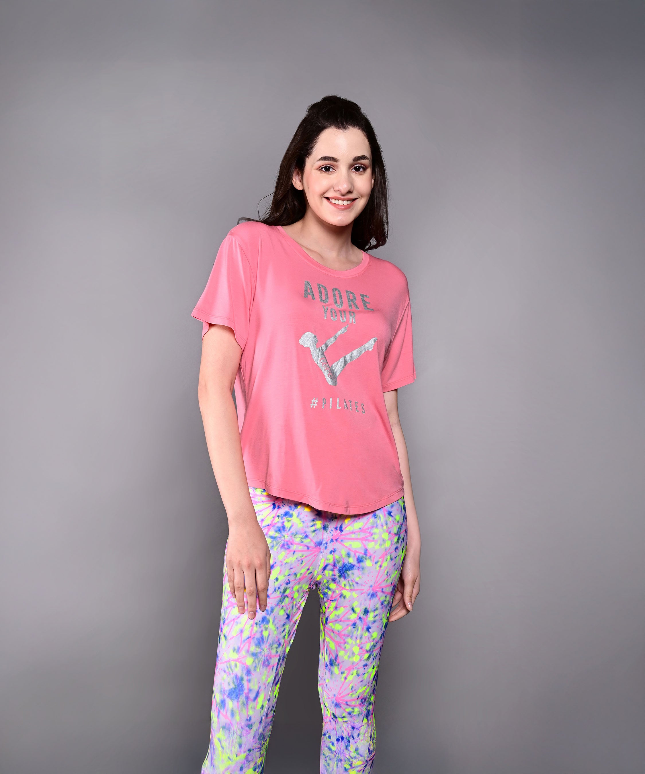 A woman in a pink 'Adore Your Core' Printed Tee smiles and poses in colorful patterned pants against a gray background—the perfect motivational shirt for any workout.