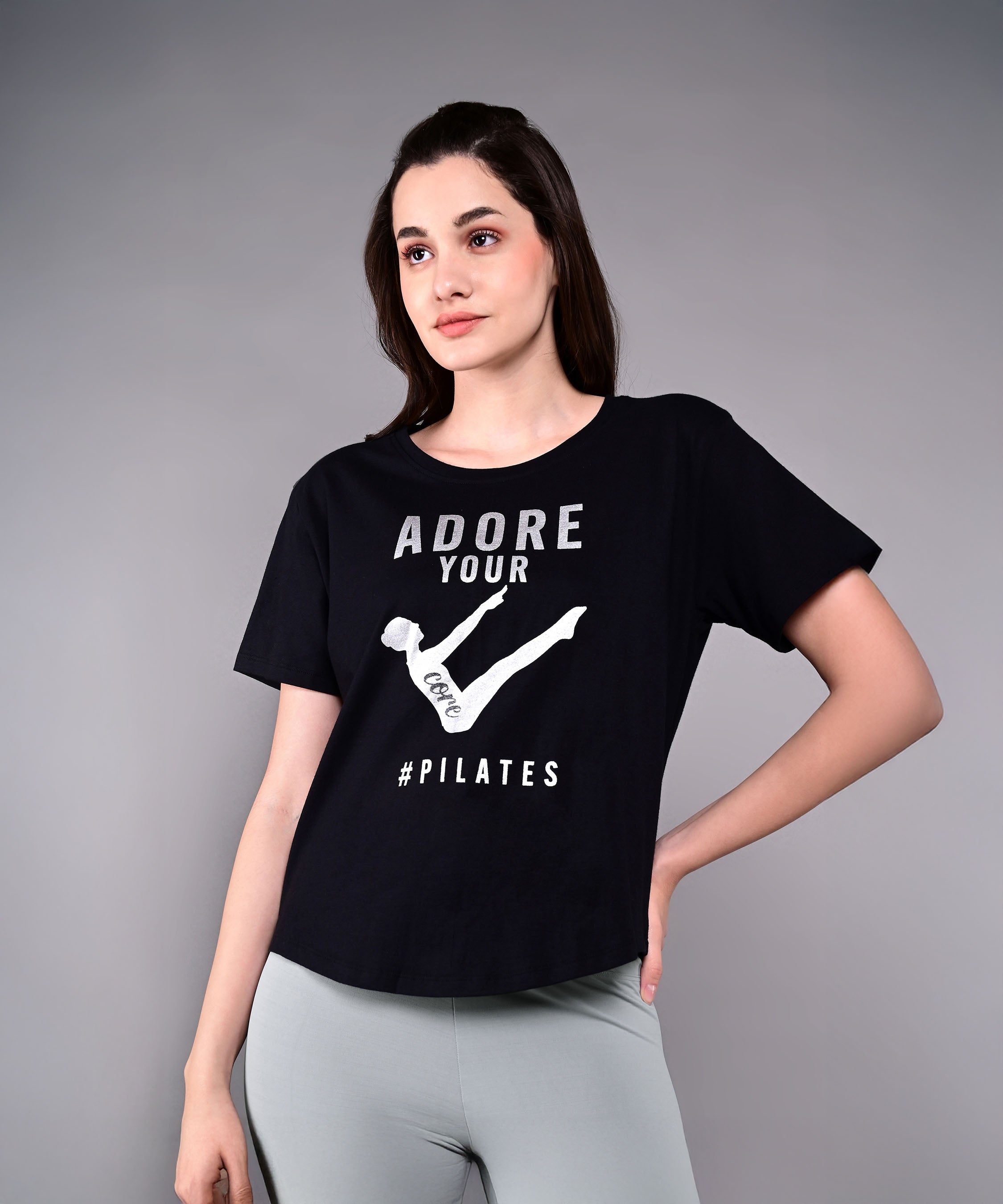 A woman wearing the 'Adore Your Core' Printed Tee and gray leggings poses against a plain backdrop, highlighting this motivational workout shirt ideal for Pilates lovers.