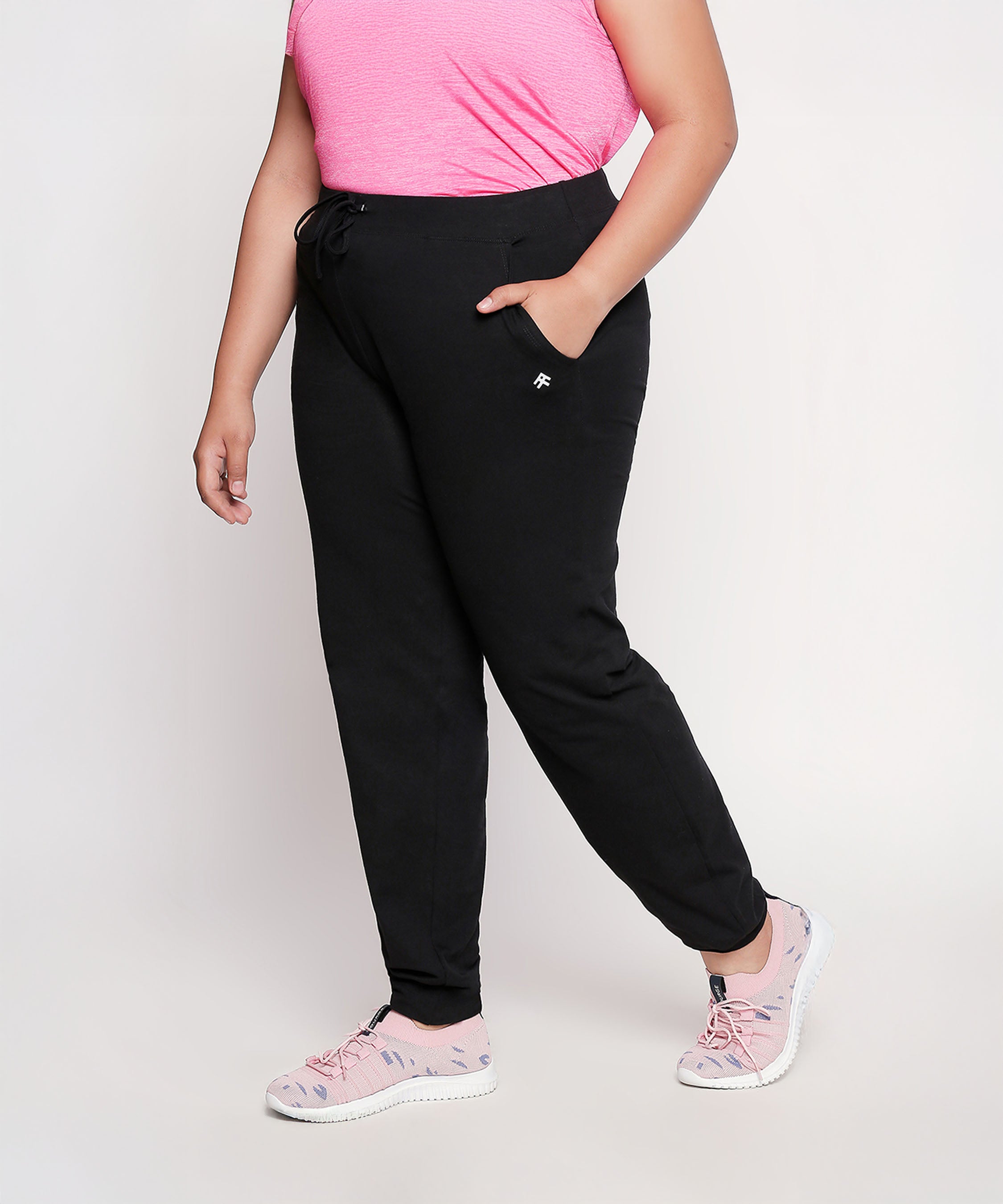 Woman in a pink top, Classic Joggers - Plus Size 02 (black high-stretchy workout pants), and pink shoes stands with one hand in her pocket.