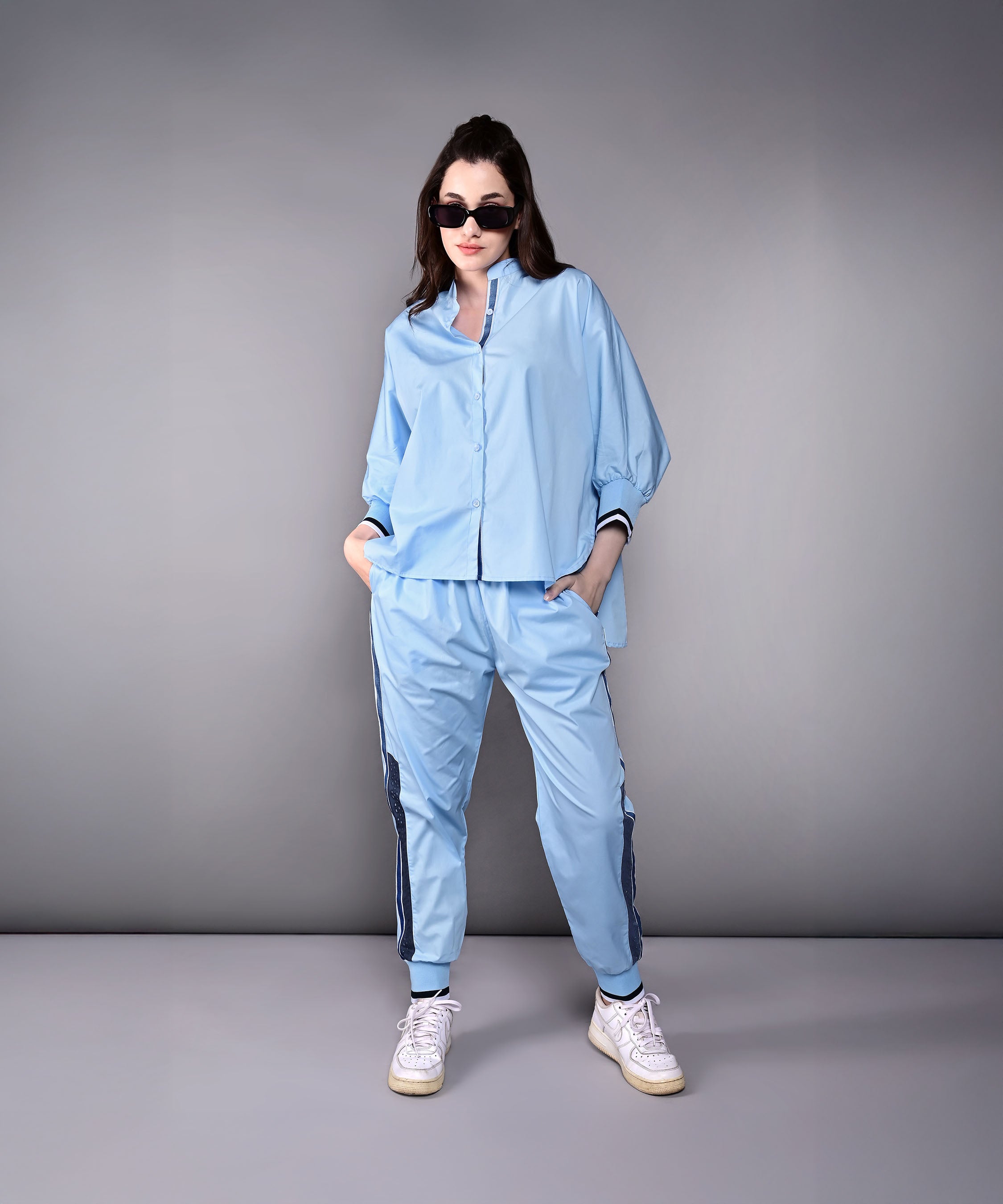 Woman wearing the Denim Accented Set—a light blue 100% cotton batwing sleeve shirt and joggers—stands with hands in pockets and sunglasses against a plain gray background.