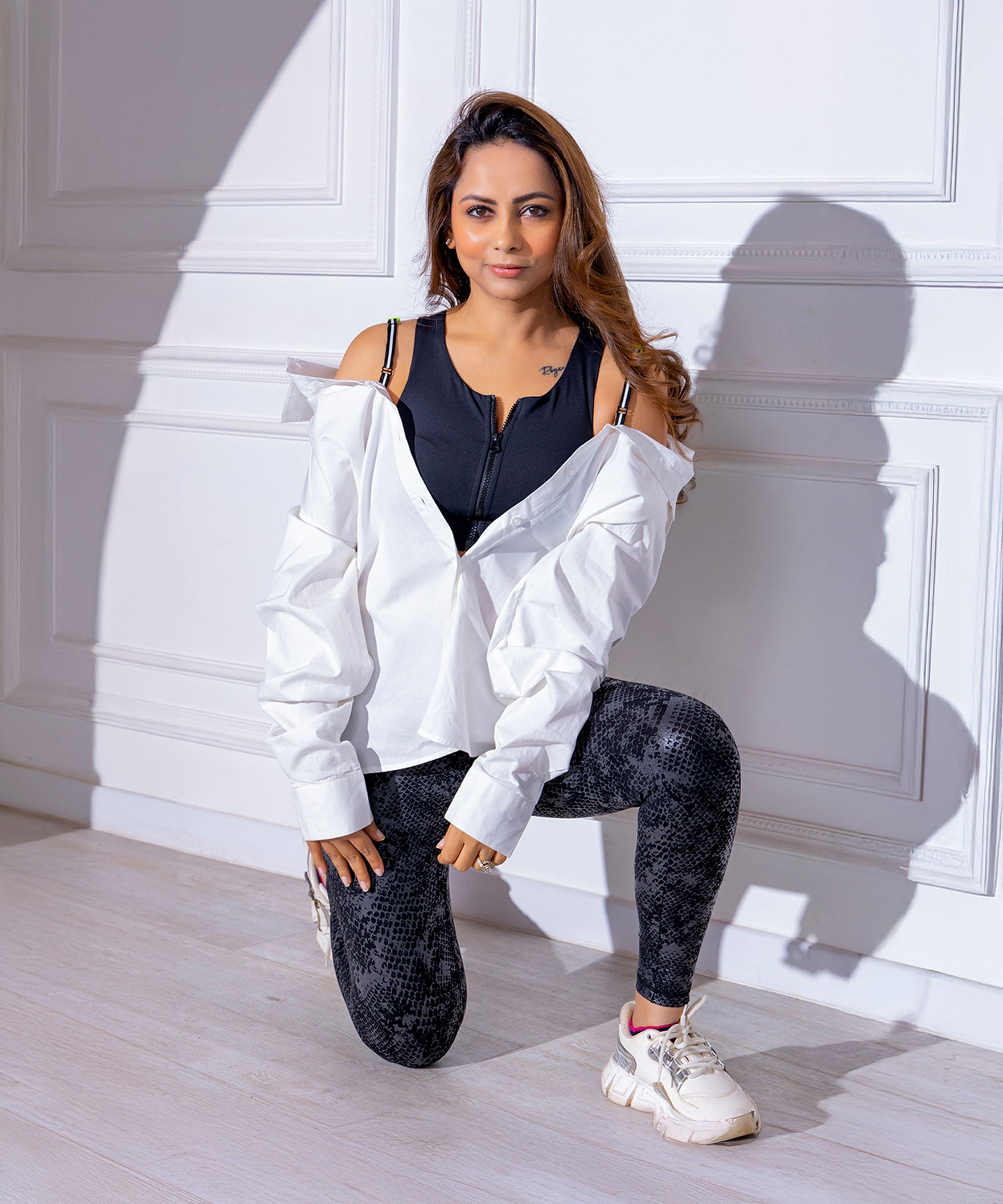 Woman kneeling indoors, wearing a White Shirt With Adjustable Straps (Free Size), patterned leggings, and white sneakers.