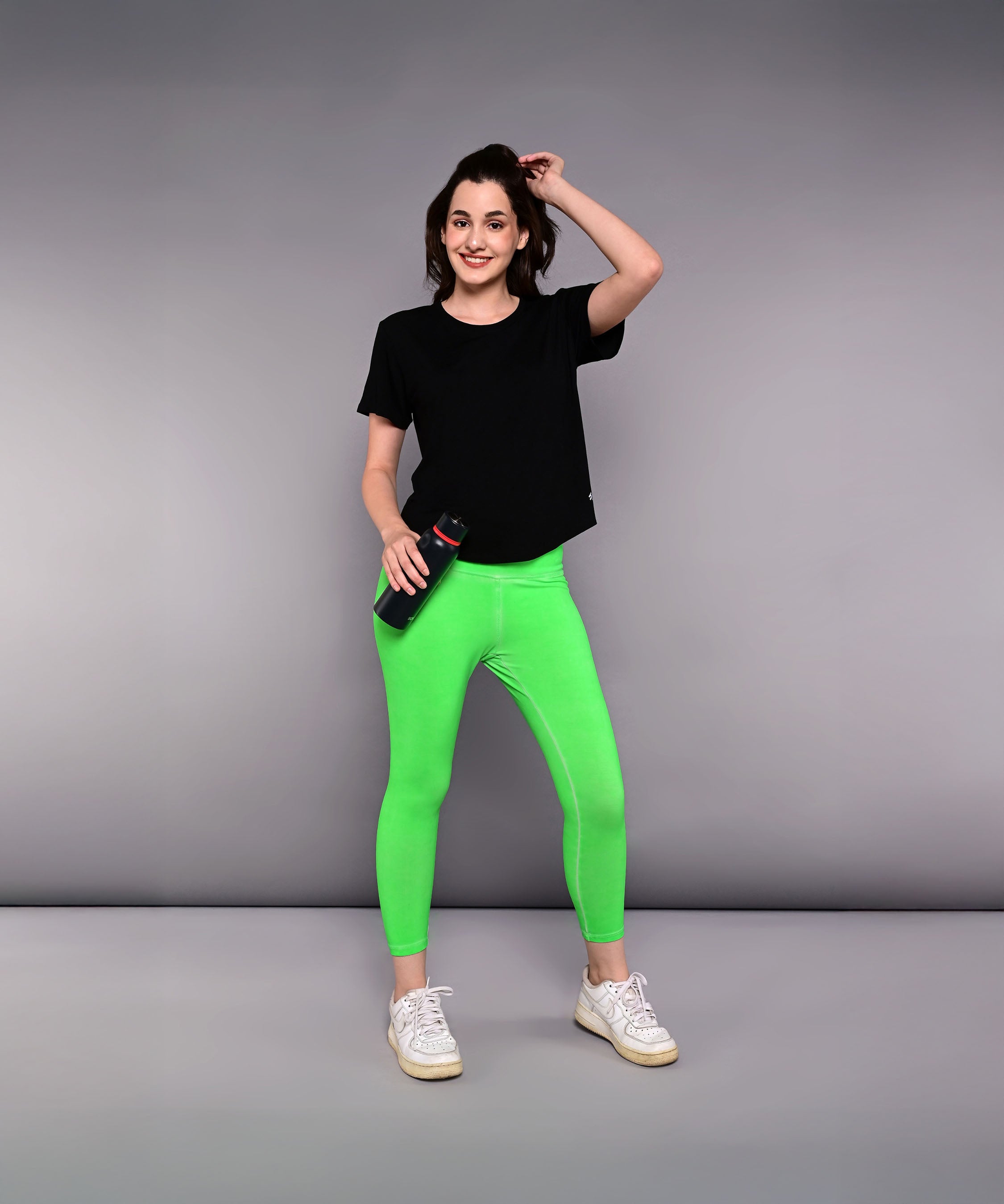 Woman in a 'Pilates Has Your Back' printed tee and neon green leggings holds a water bottle, standing against a plain gray background.
