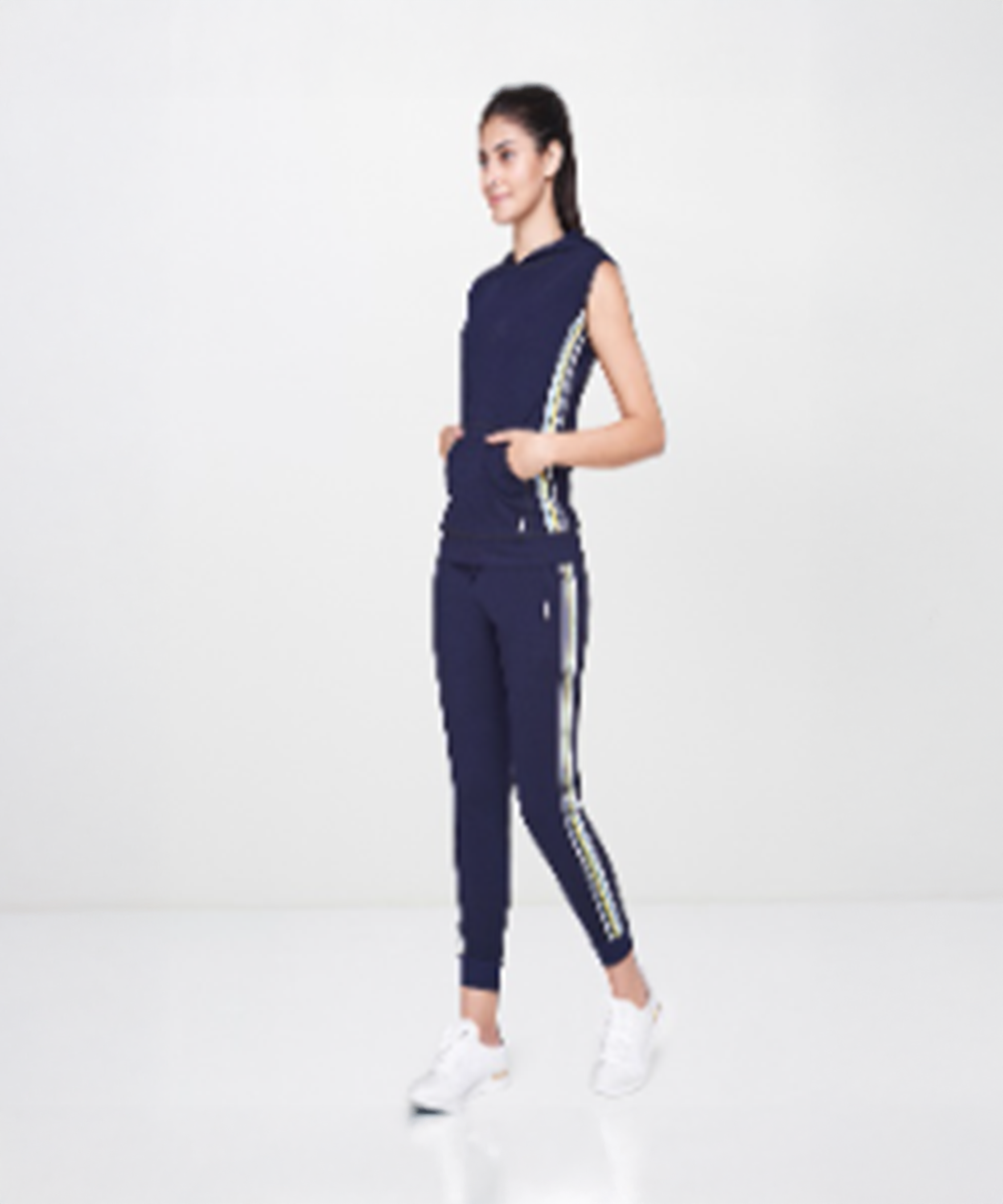 A woman stands in a white studio wearing a Navy Blue Sleeveless Hoodie and matching navy track pants with side stripes.