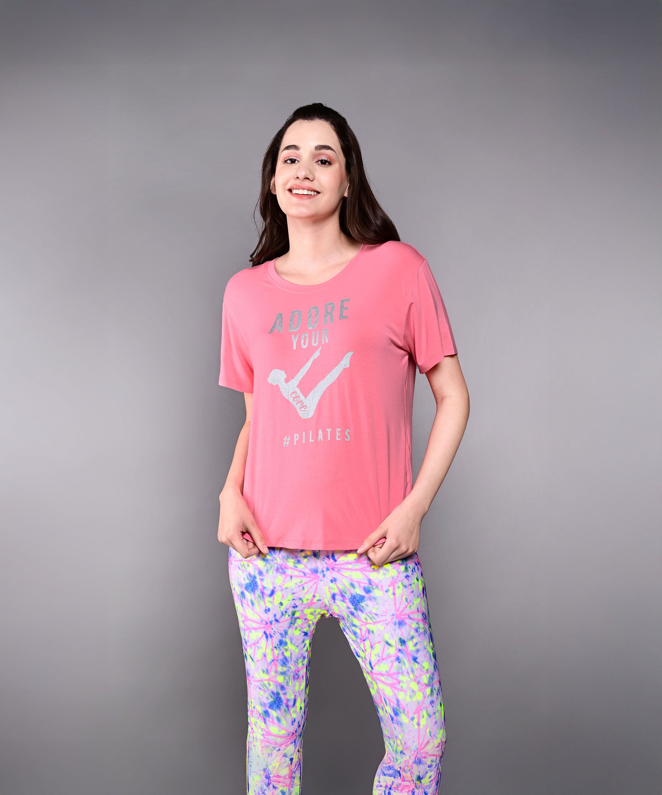A woman smiles, wearing the 'Adore Your Core' Printed Tee in pink with vibrant patterned leggings against a gray background. This motivational shirt adds a touch of positivity to any fitness routine.