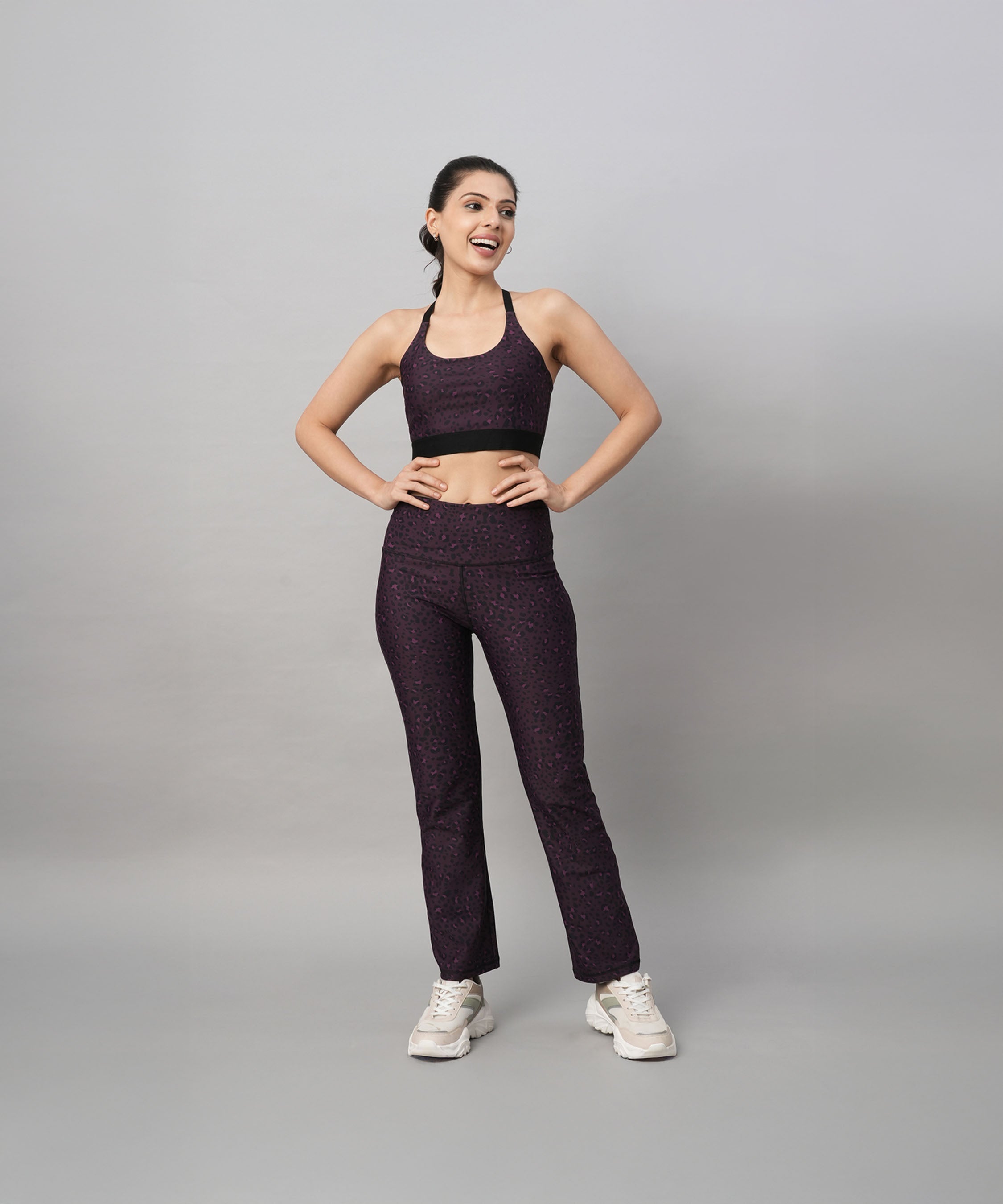 A woman in matching dark workout clothes stands smiling with hands on hips, highlighting the Pink Animal Print Sports Bra against a plain background.