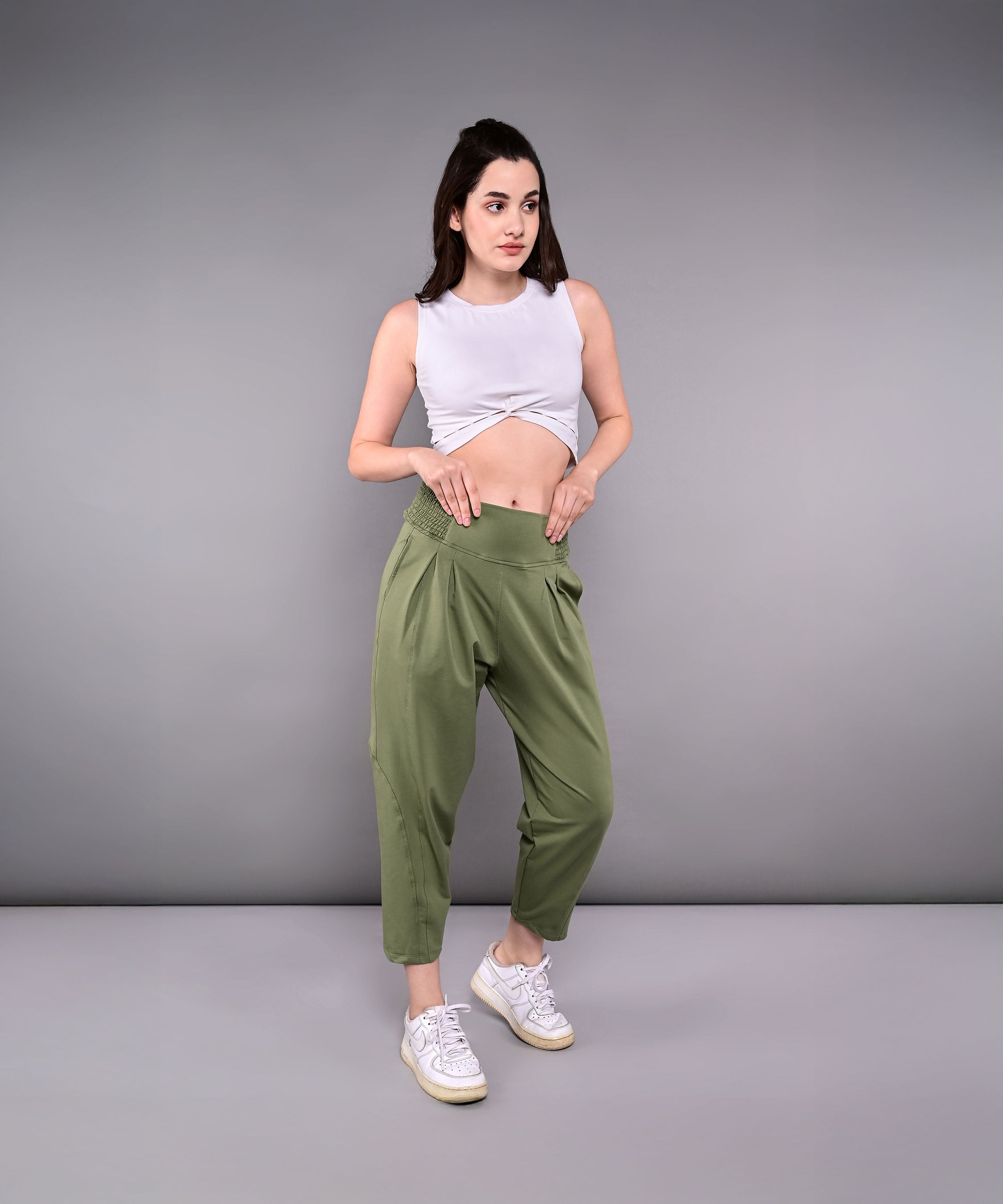 Woman indoors wearing Pleated Yoga Pant With Smocked Waistband in olive green, paired with a white sleeveless crop top and white sneakers.