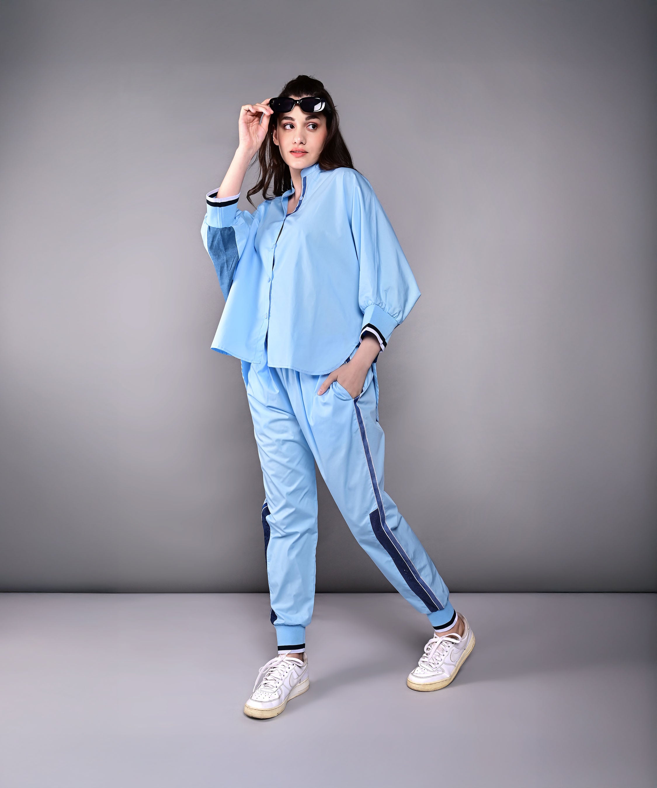 Woman models the Denim Accented Set—light blue shirt and joggers paired with white sneakers—against a plain gray background, highlighting the comfort of 100% cotton.