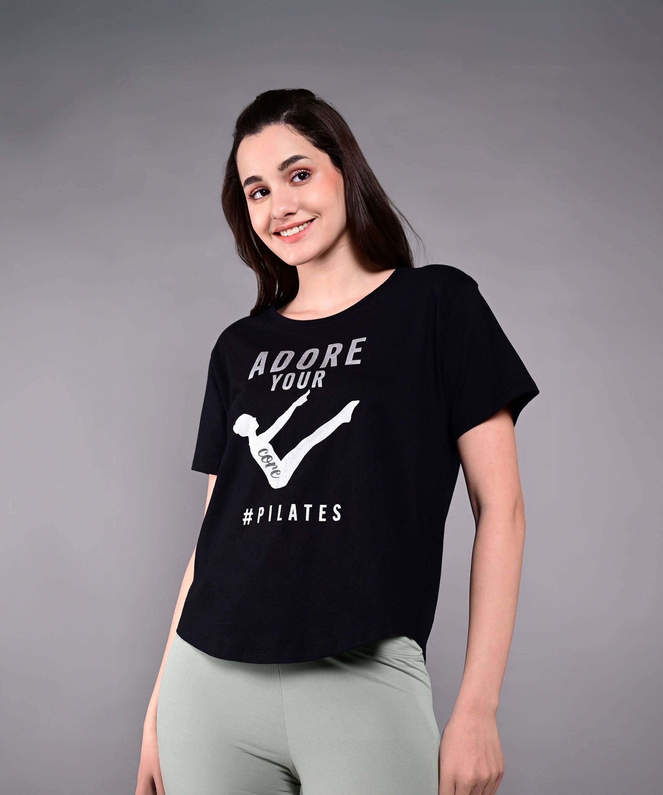 Smiling woman in light leggings, wearing our 'Adore Your Core' Printed Tee against a grey backdrop. This women's cotton modal shirt brings comfort and style—ideal for any Pilates fan.