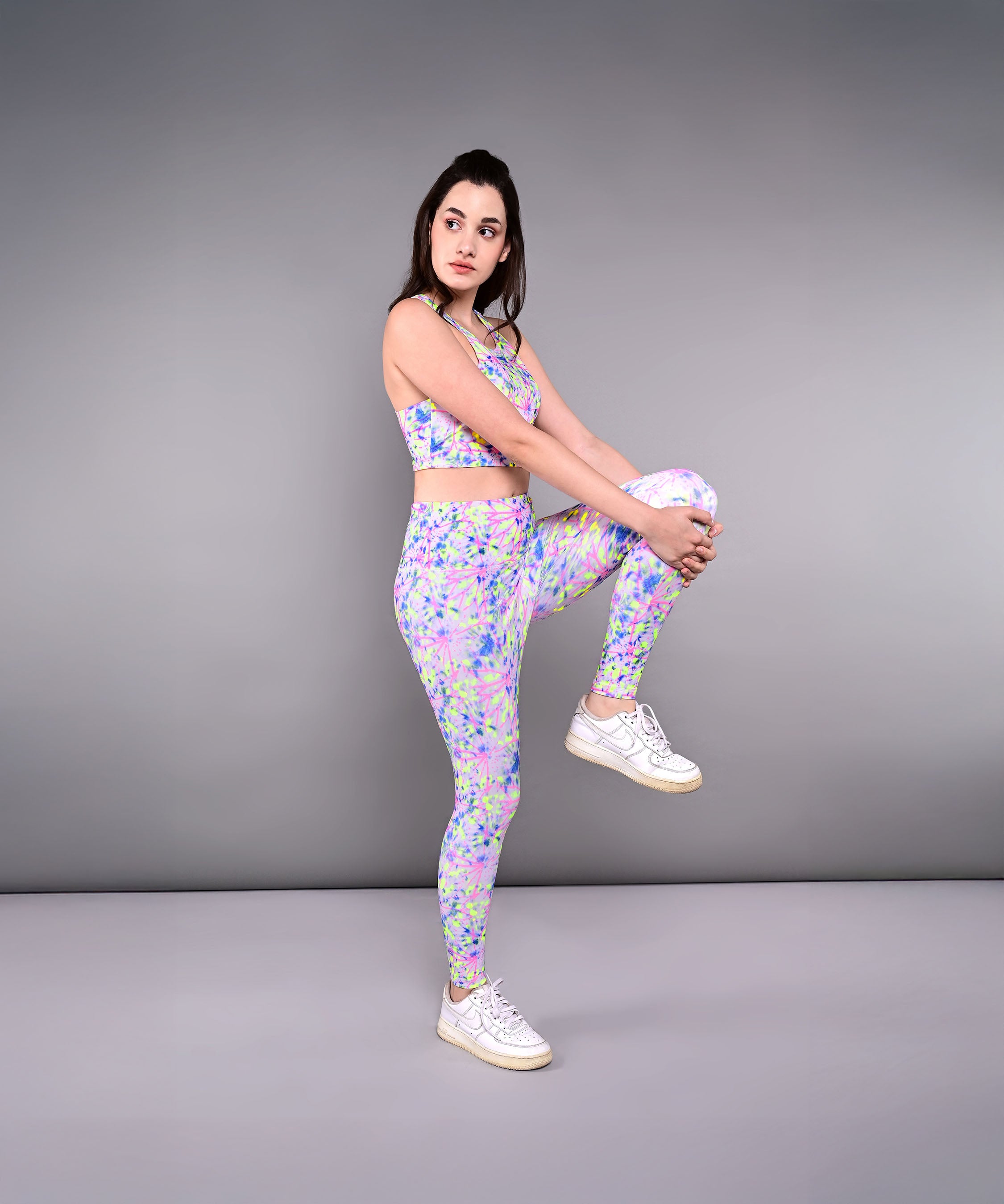 A woman wears the Multicolor Neon Crop Top, made from skin-friendly cotton lycra, as she performs a one-legged yoga pose against a plain gray background.