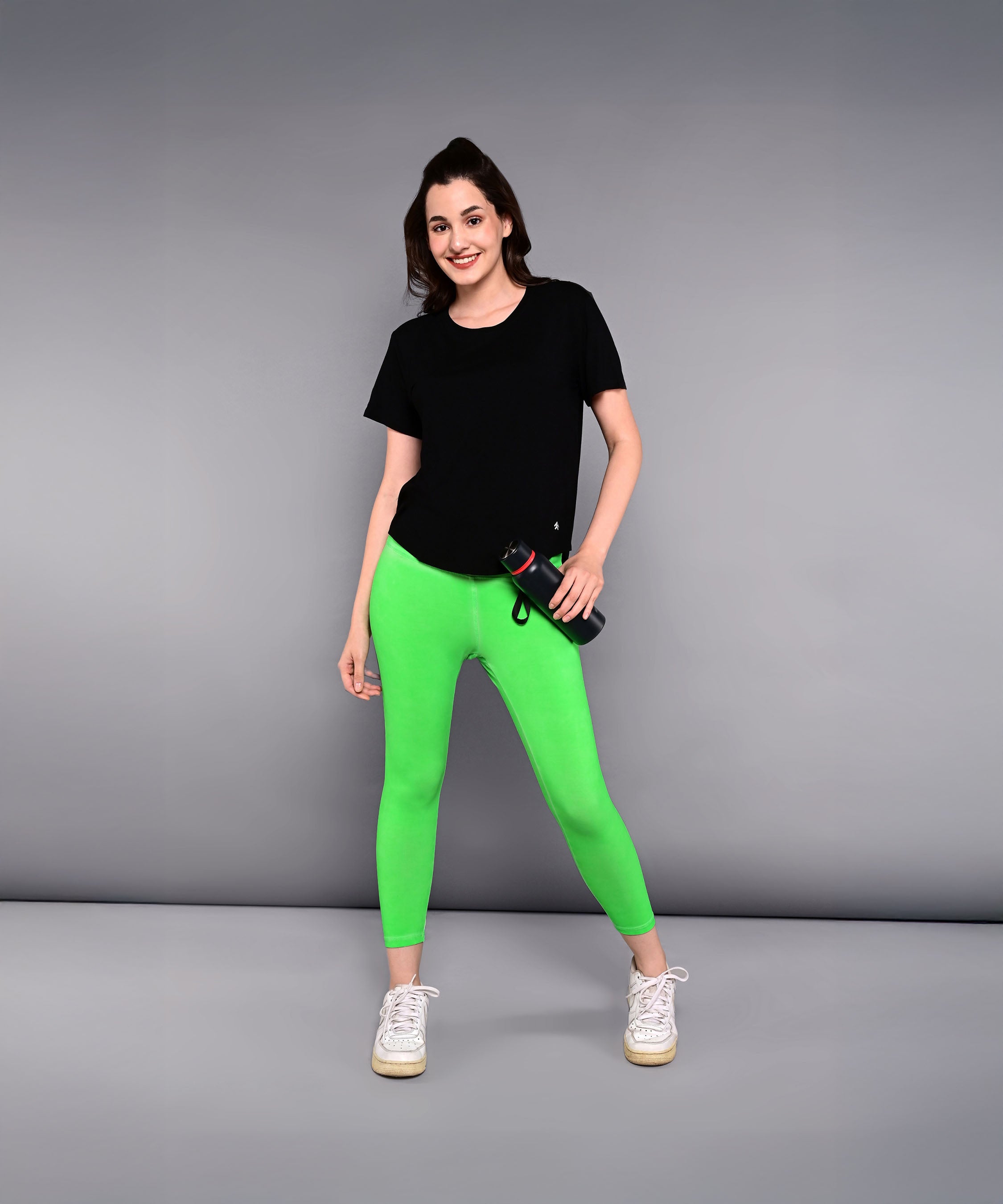 Woman in a 'Pilates Has Your Back' Printed Tee and bright green leggings, holding a water bottle and standing against a gray background.