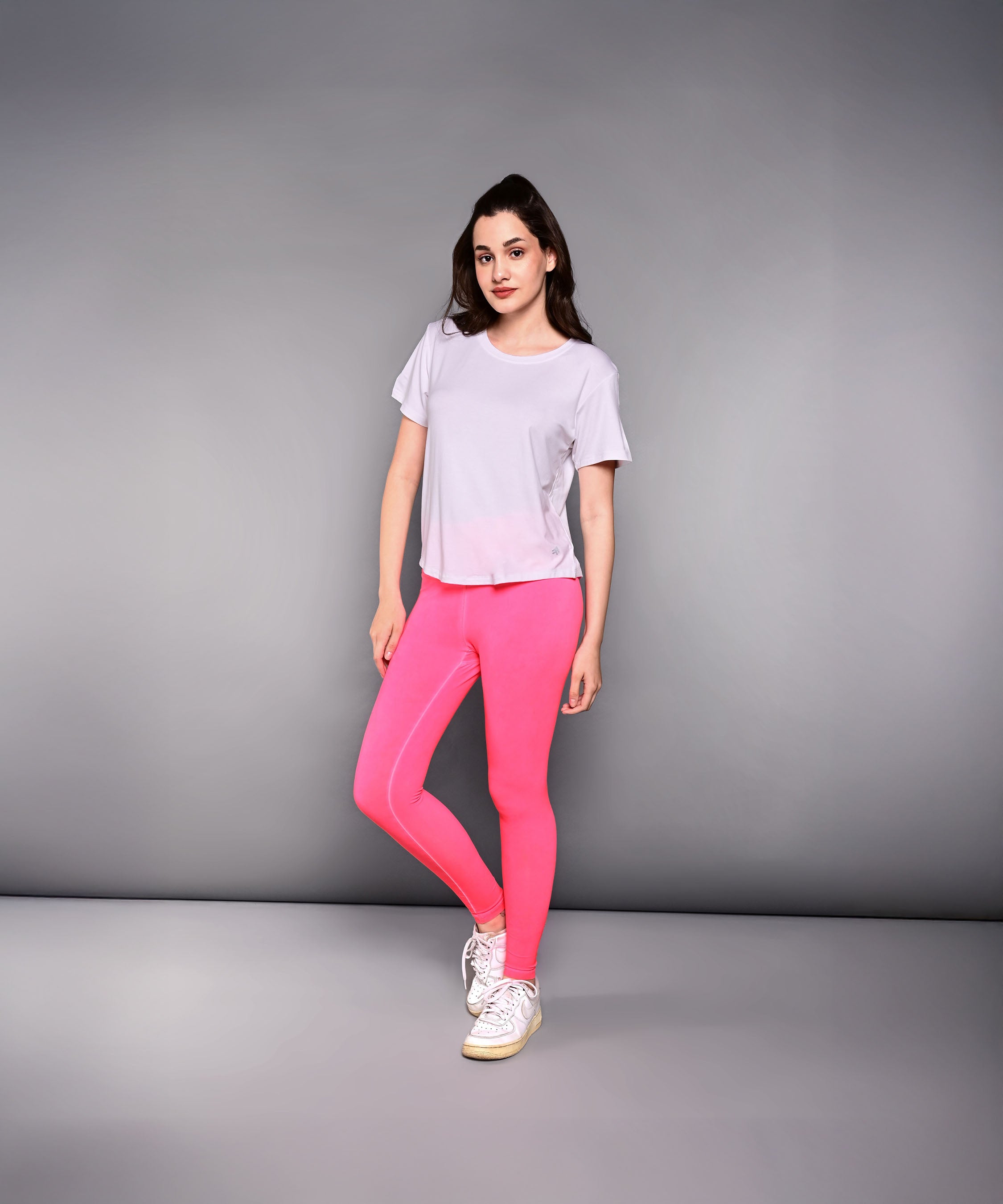 Woman stands in pink leggings, 'Pilates Has Your Back' printed tee, and white sneakers against a plain gray background.