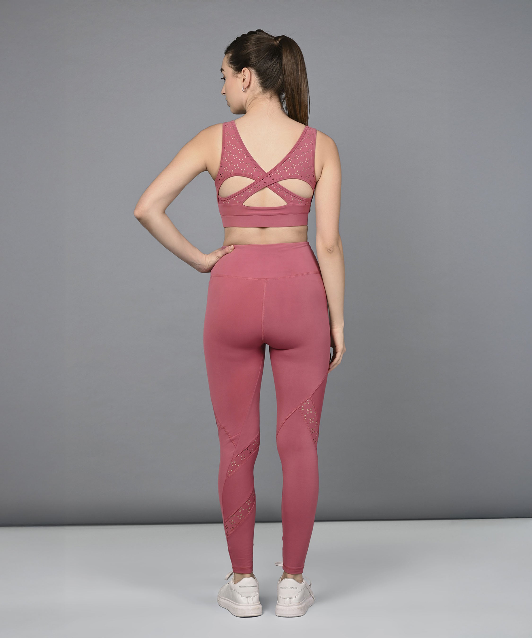A woman stands with her back to the camera, wearing a Peony Pink Netted Top featuring a HoneyComb Self Pattern and matching leggings.