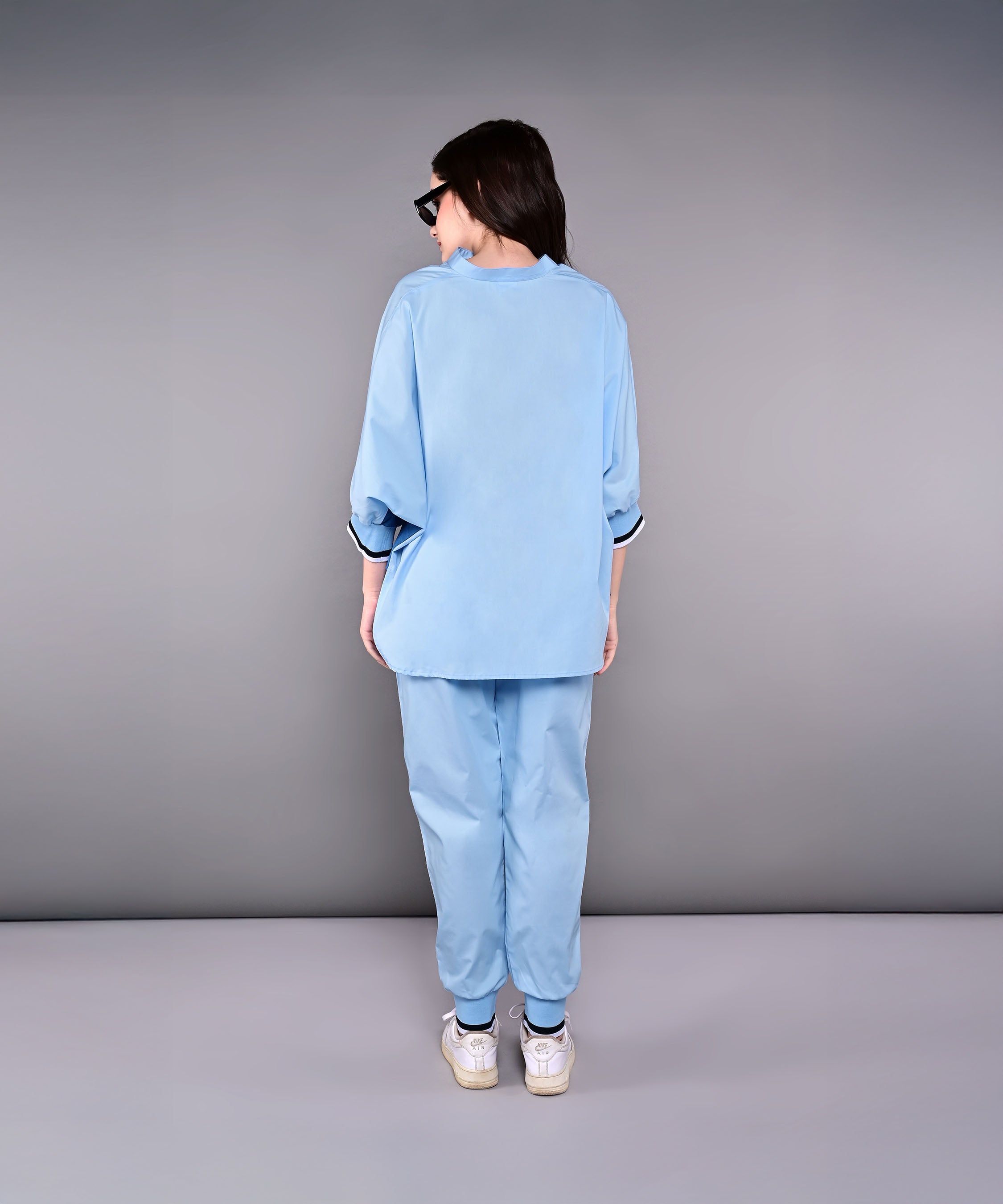 Woman wearing the Denim Accented Set—loose 100% cotton batwing sleeve shirt and joggers—with white sneakers, standing with her back to the camera against a gray backdrop.