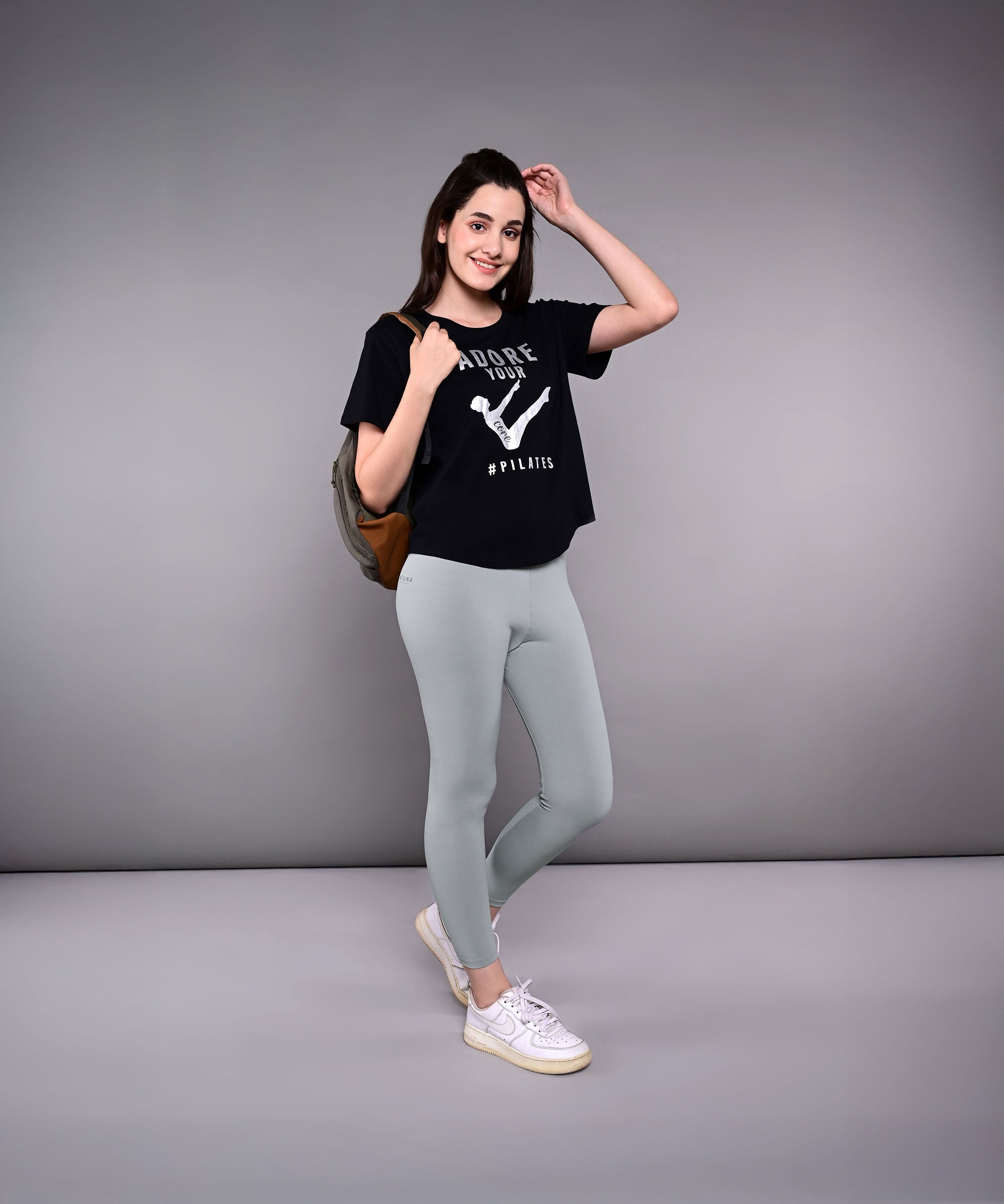Woman wearing the 'Adore Your Core' Printed Tee with gray leggings, a backpack, and white sneakers poses and smiles.