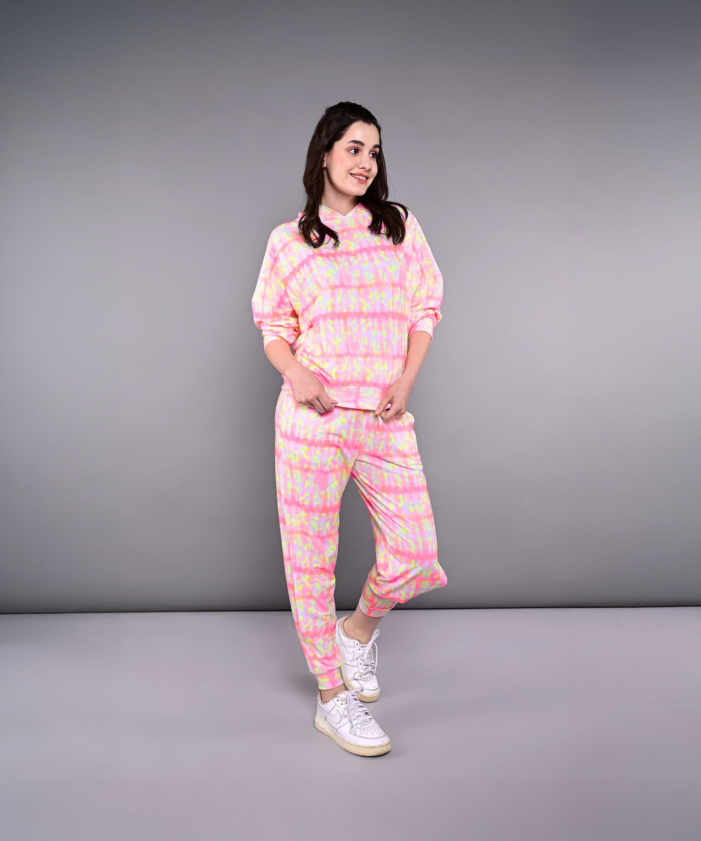 Woman in the Comfort Fit Tie-Dye Sweatshirt With Joggers Set, featuring a pink and yellow tie-dye hooded sweatshirt and joggers in breathable poly-blend fabric, paired with white sneakers, smiling against a gray background.
