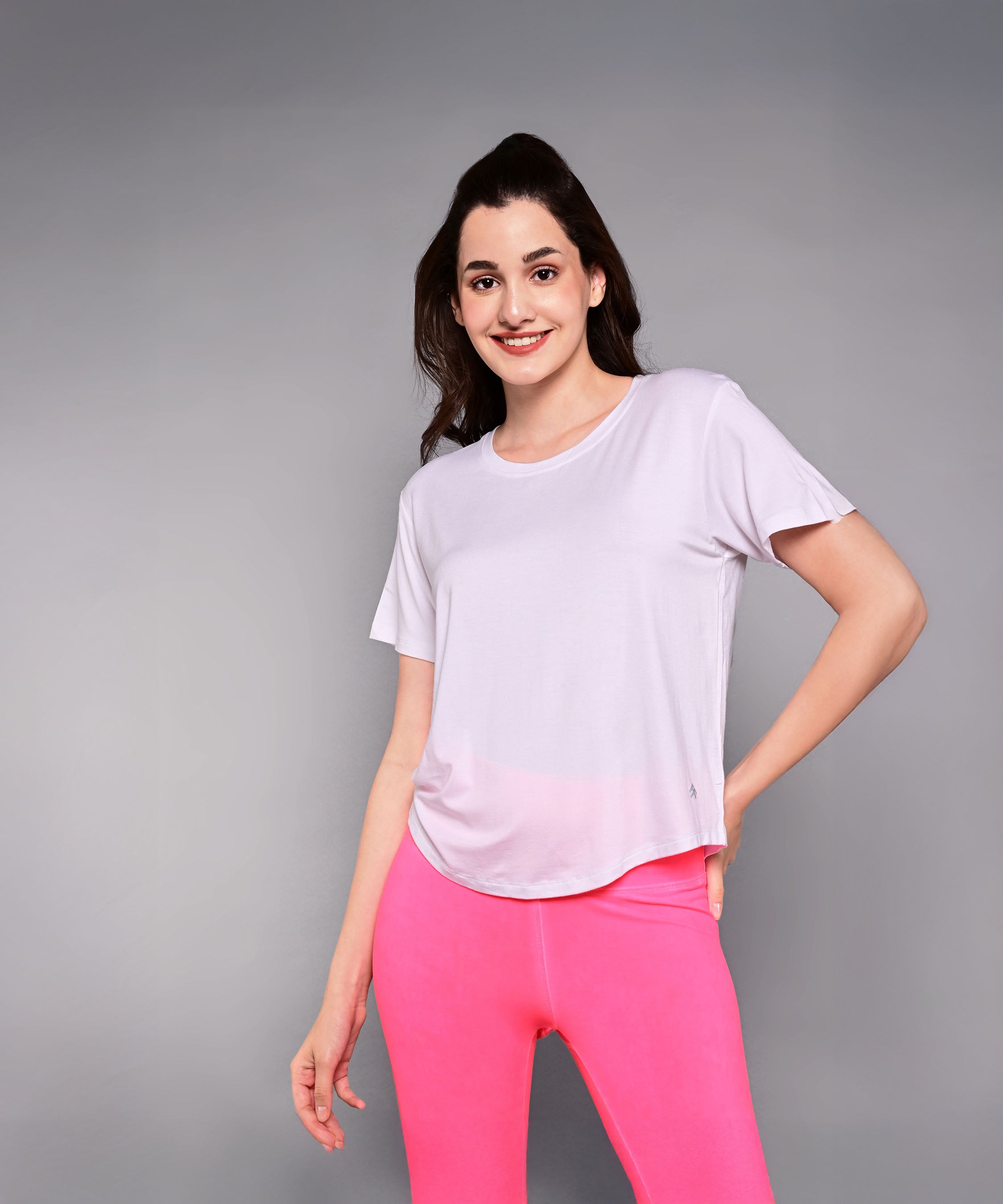A woman in a light pink 'Pilates Has Your Back' Printed Tee and bright pink leggings smiles confidently against a gray background—the ideal motivational shirt for any Pilates tee collection.