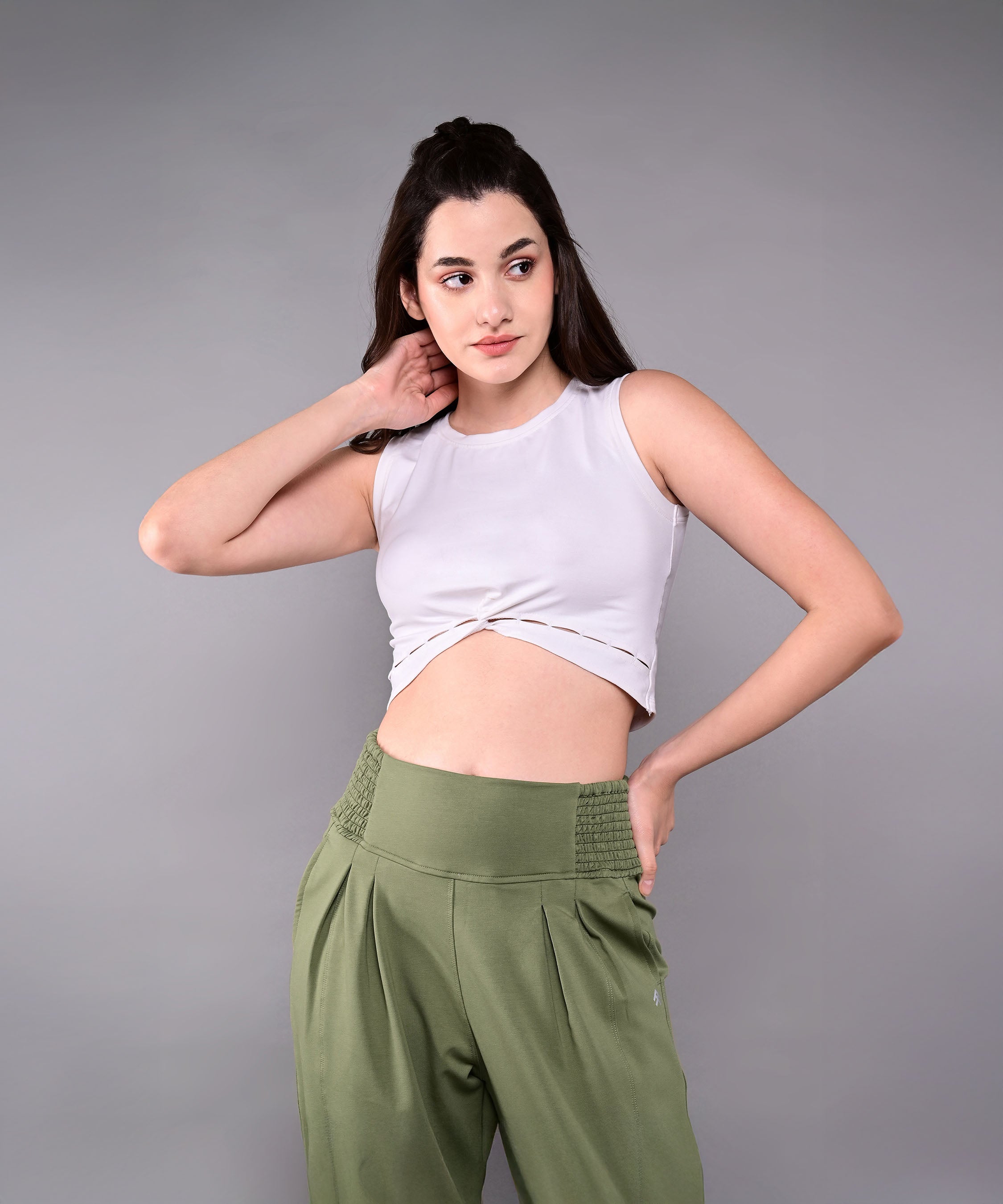 A woman poses against a gray background wearing a sleeveless white crop top and Pleated Yoga Pant With Smocked Waistband in green, with one hand behind her head.