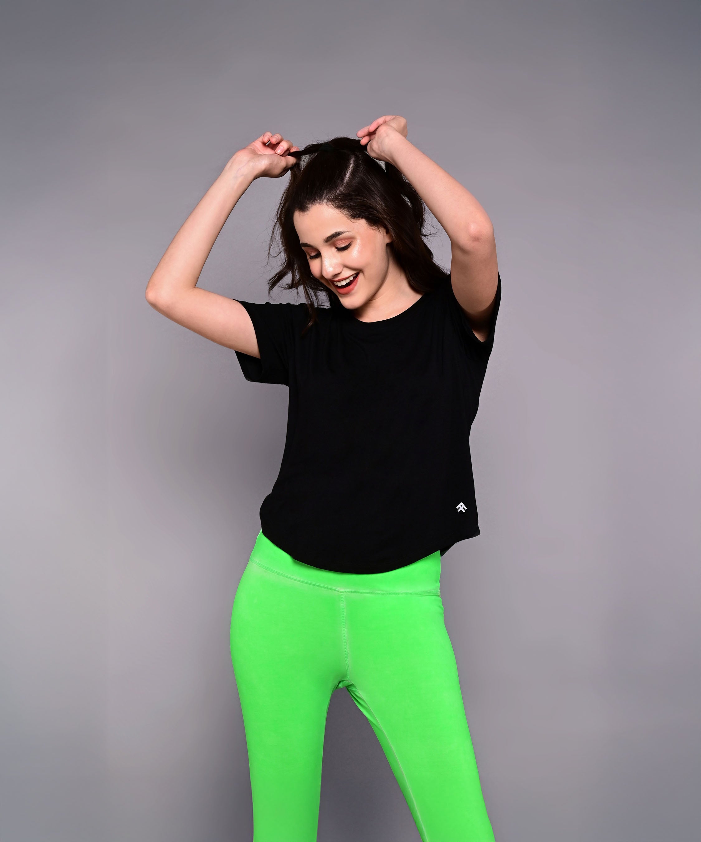A woman smiles with hands raised, wearing the 'Pilates Has Your Back' Printed Tee and bright green leggings against a gray background.