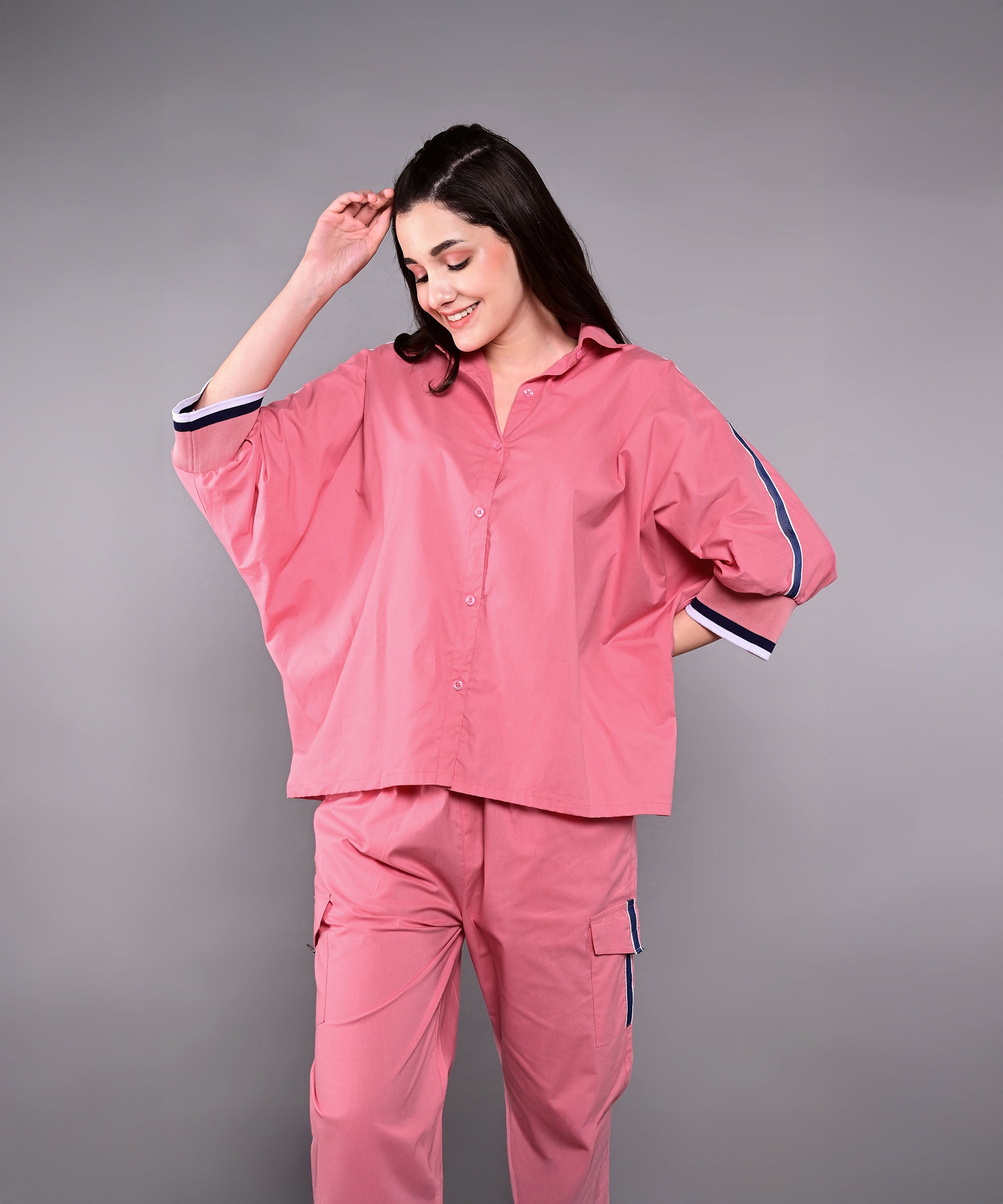 A woman smiles in the Effortlessly Chic Set—a loose pink cotton co-ord with striped batwing sleeve shirt and cargo track pants—posing against a plain gray background.