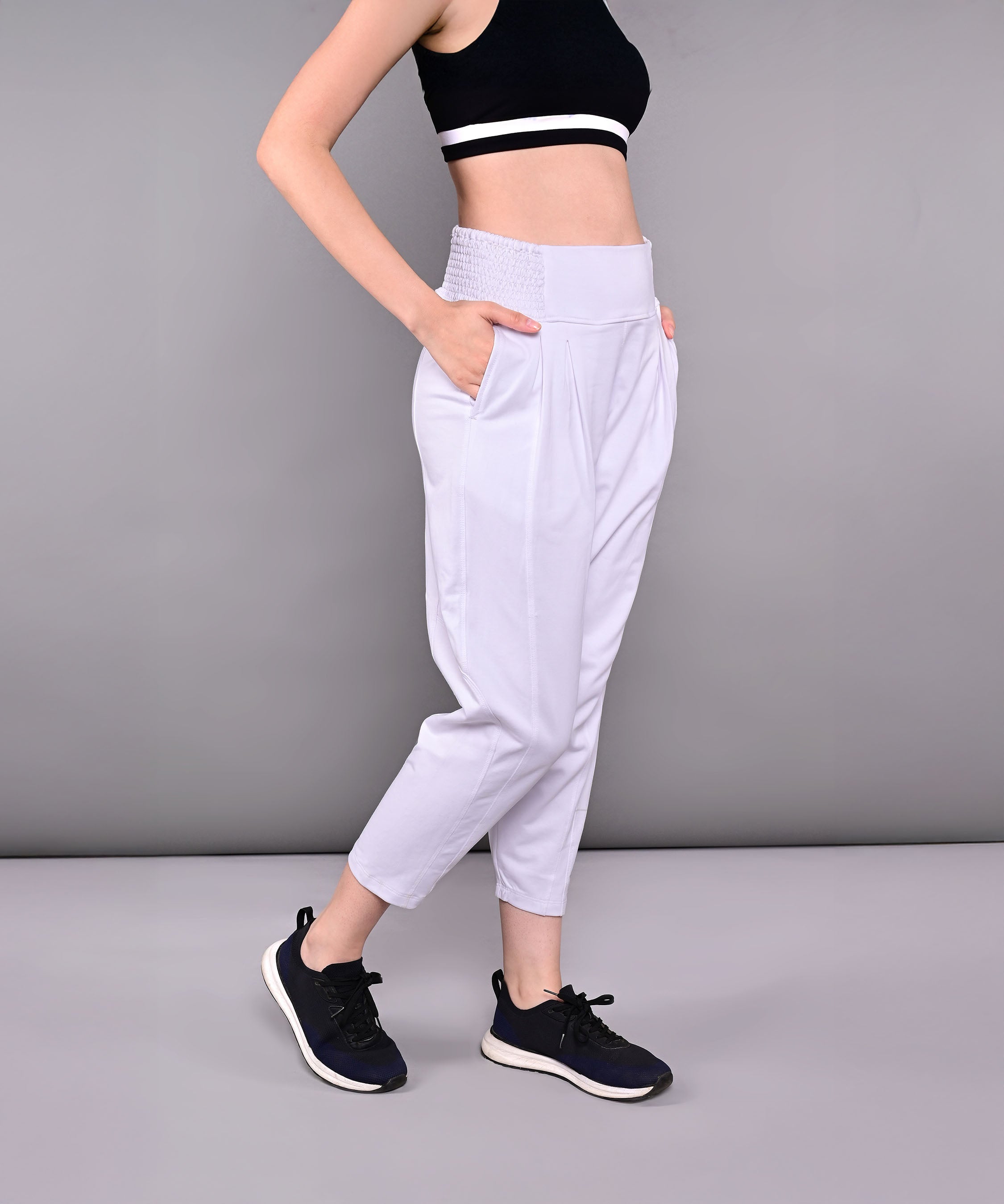 Woman wearing Pleated Yoga Pant With Smocked Waistband in light gray cotton, paired with a black crop top, hands in pockets, standing against a gray background.