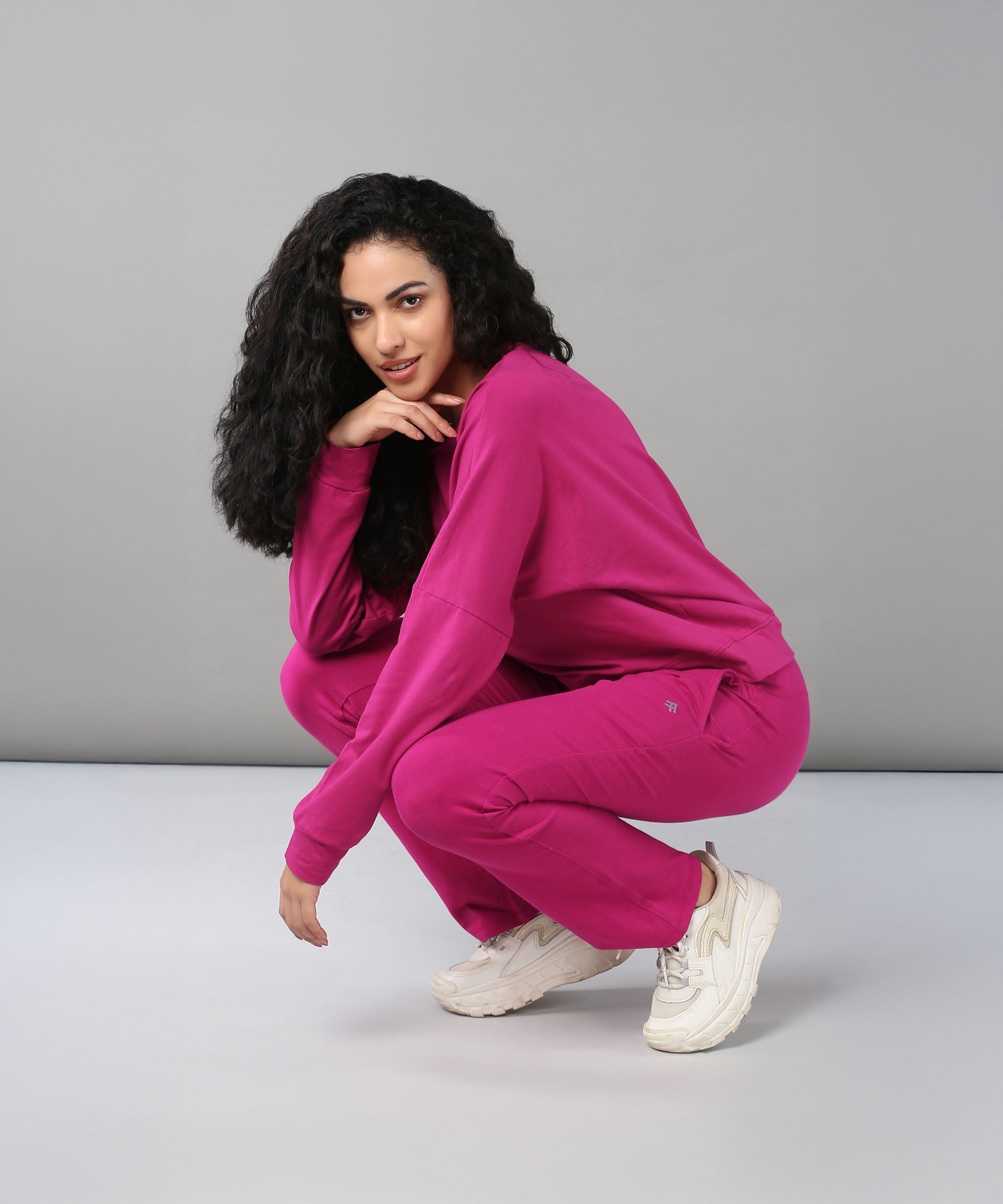 A woman with curly hair squats in the Deep Pink Comfort Duo, pairing a cotton lycra top and matching joggers with white sneakers, set against a gray background.