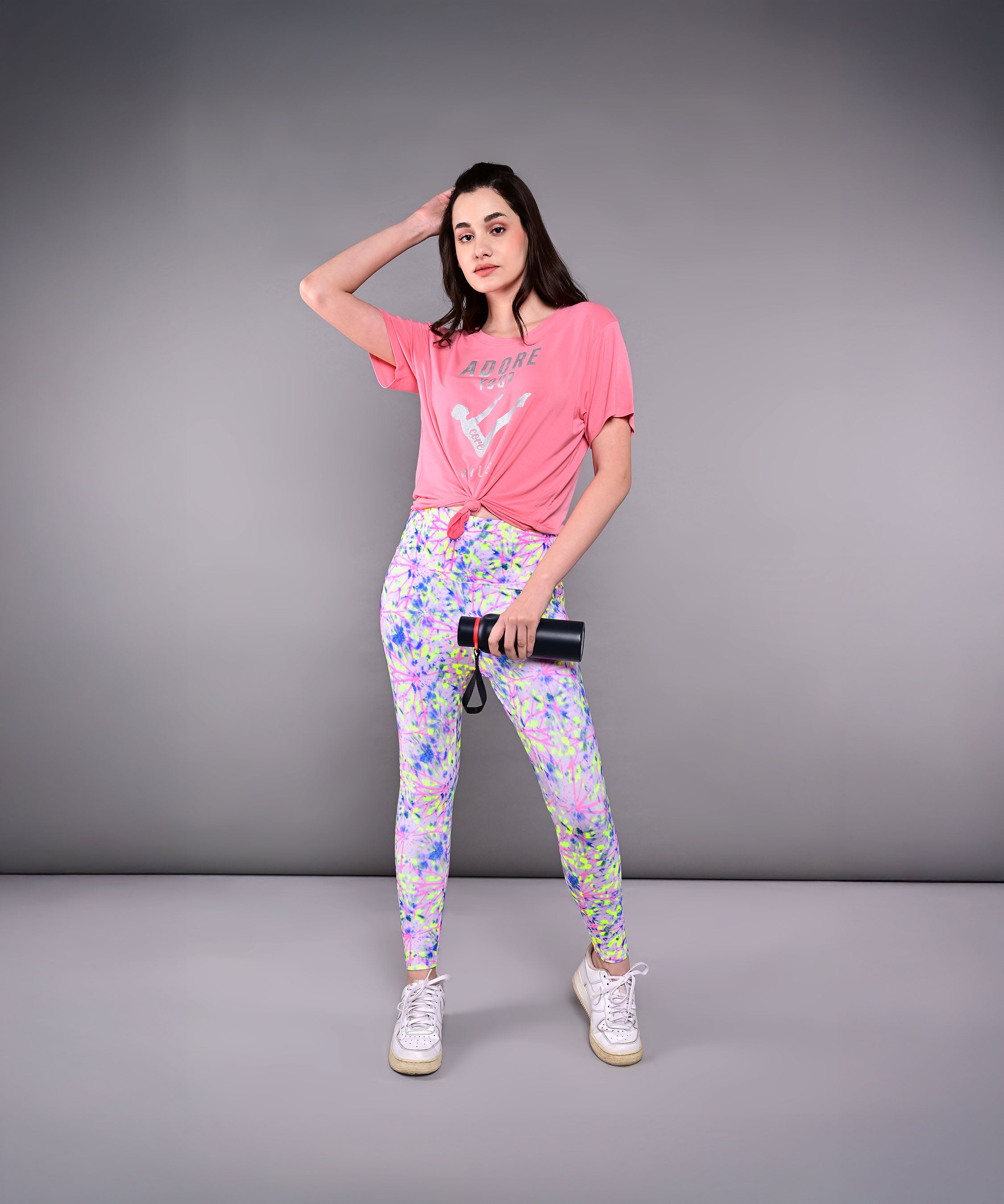 A woman poses against a gray background, holding a camera and wearing the 'Adore Your Core' Printed Tee with colorful leggings—her look radiates energy and confidence.