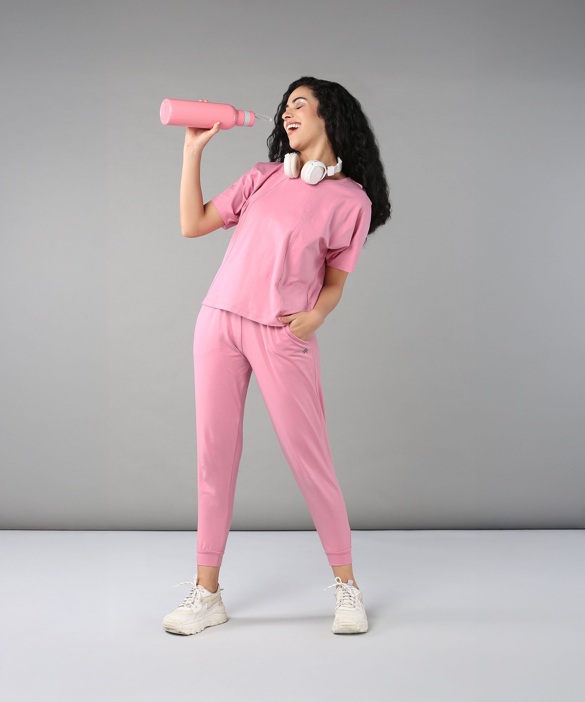 Wearing the Flamingo Pink Chic Ensemble—joggers in buttery soft fabric and a racerback top—a woman with headphones around her neck smiles, holding a pink water bottle against a gray background.