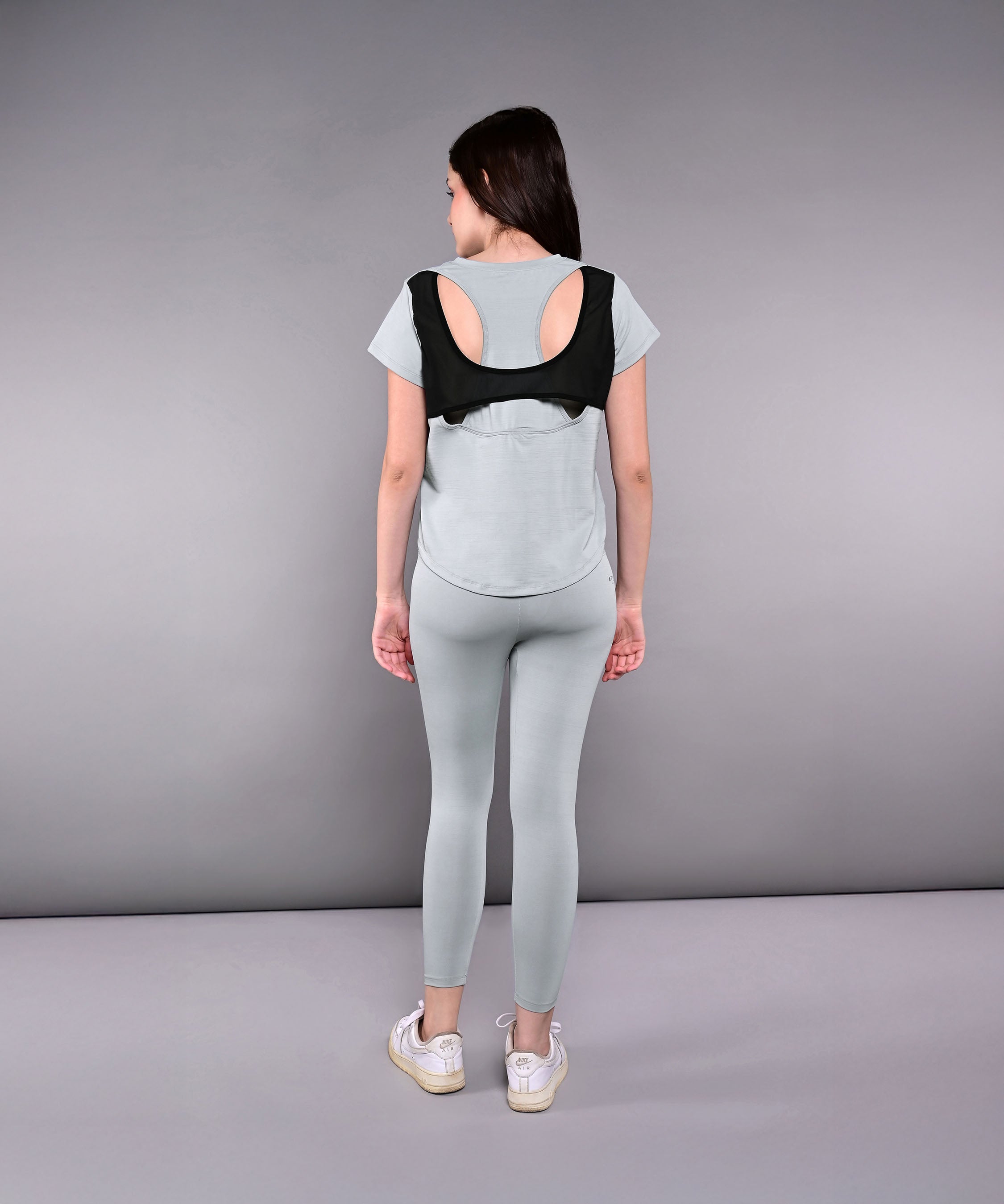 A woman in the Dynamic Grey Performance Set—light gray athletic leggings and a racerback top—faces away, wearing a black posture corrector over her workout outfit.