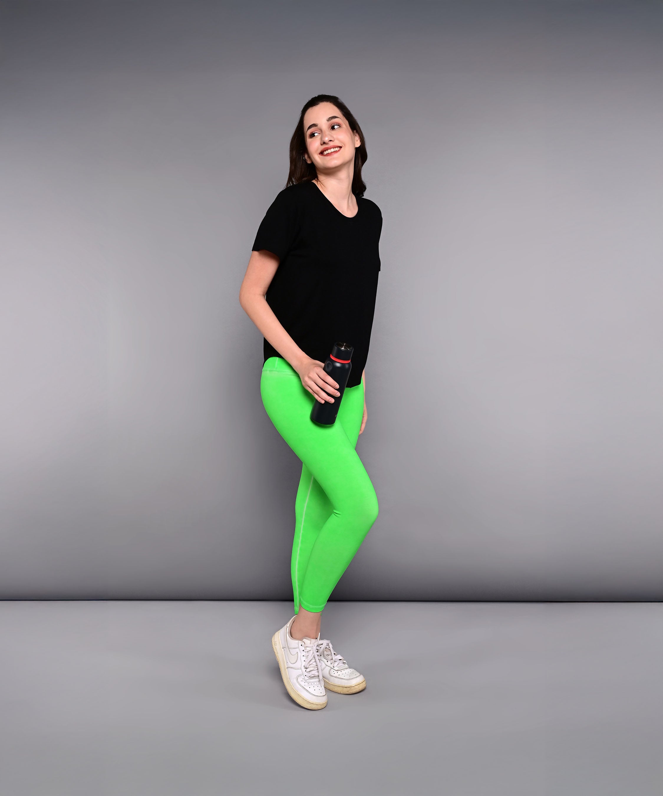 Woman in a 'Pilates Has Your Back' printed tee and bright green leggings holding a water bottle, standing against a gray background.