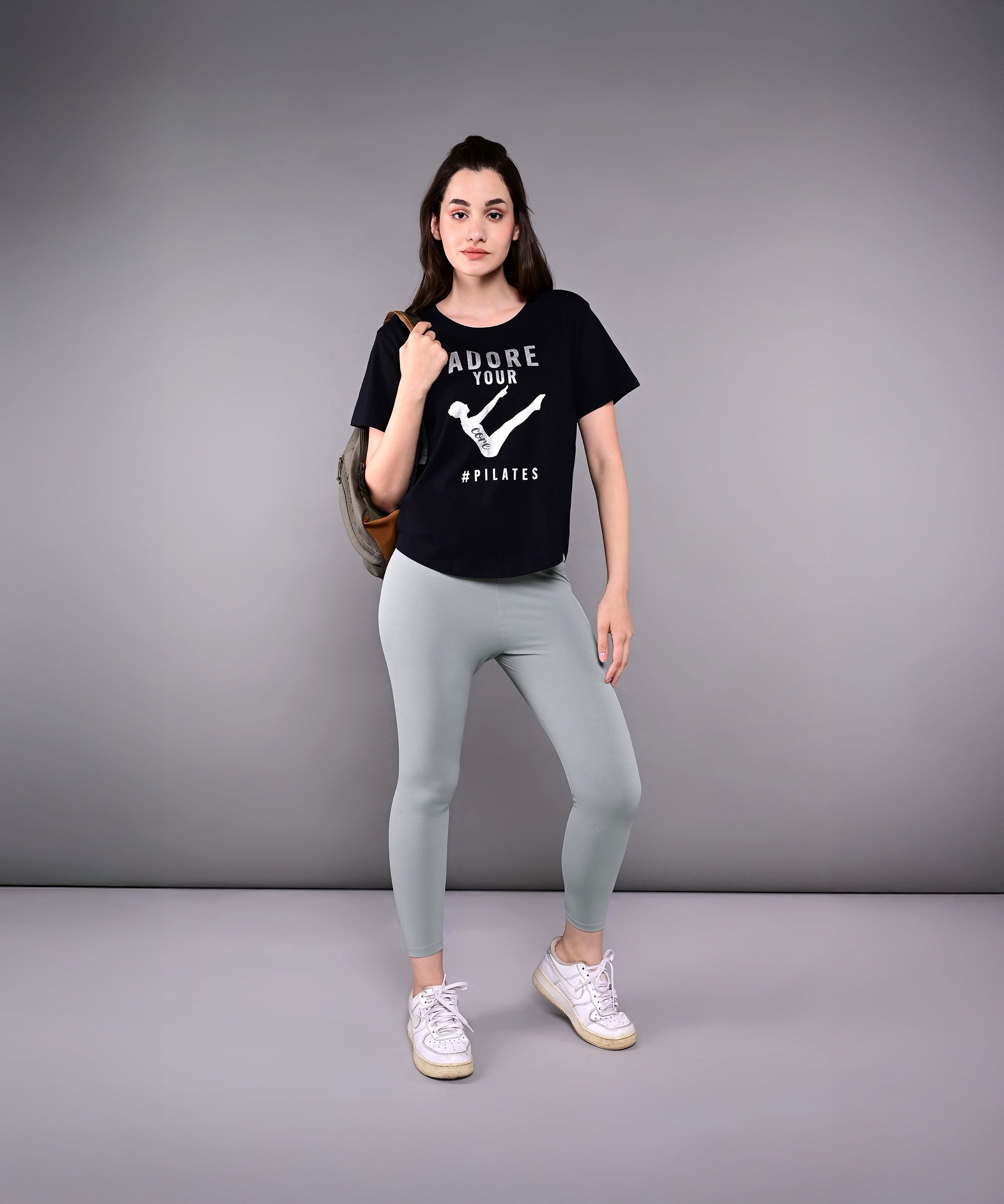 A woman stands confidently in our 'Adore Your Core' Printed Tee, pairing the black cotton-modal shirt with gray leggings and sneakers against a plain background.