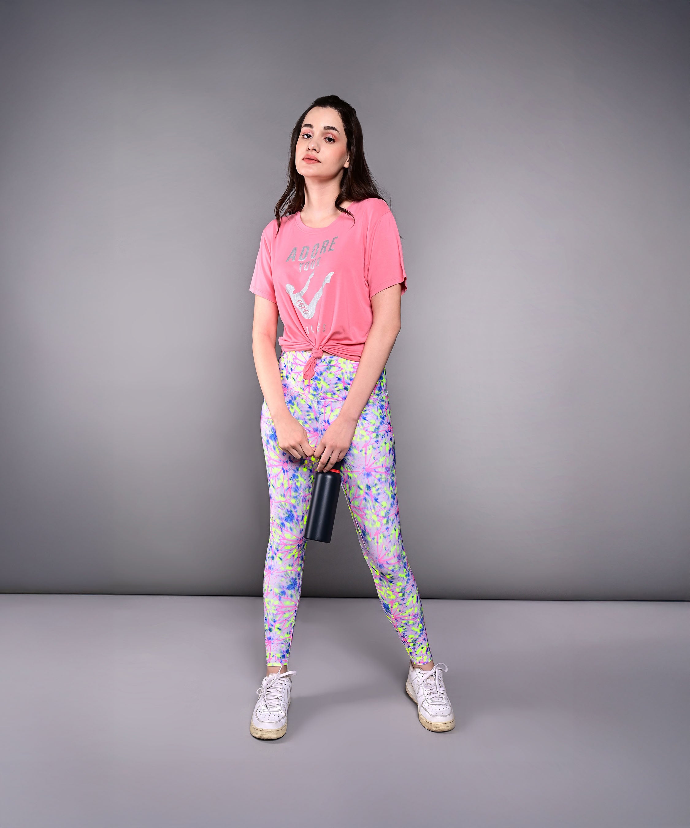 Young woman in the 'Adore Your Core' Printed Tee and patterned leggings holds a tumbler while standing against a plain gray background.