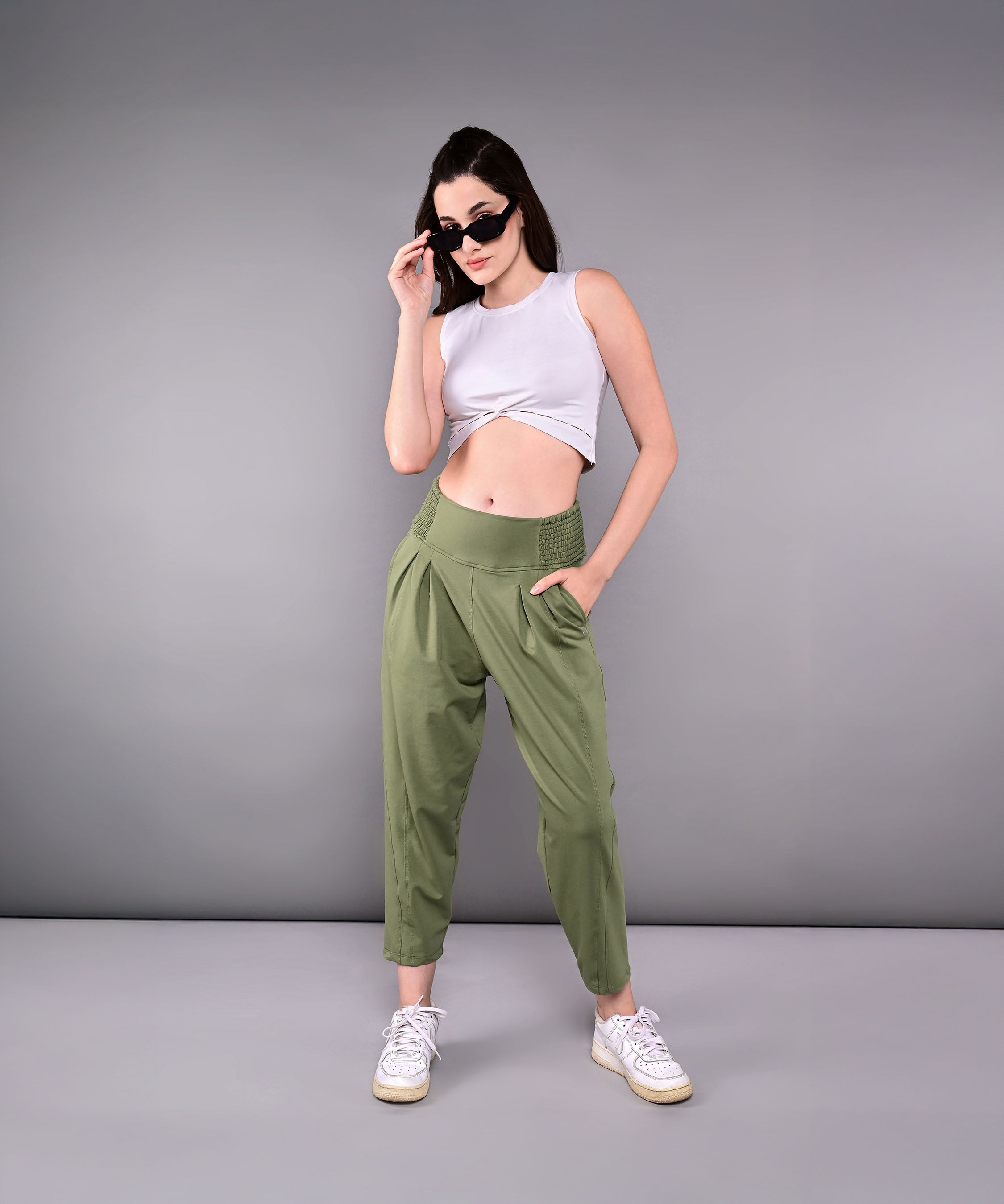 A woman in sunglasses and a white crop top showcases the Pleated Yoga Pant With Smocked Waistband, posing confidently against a plain gray background.