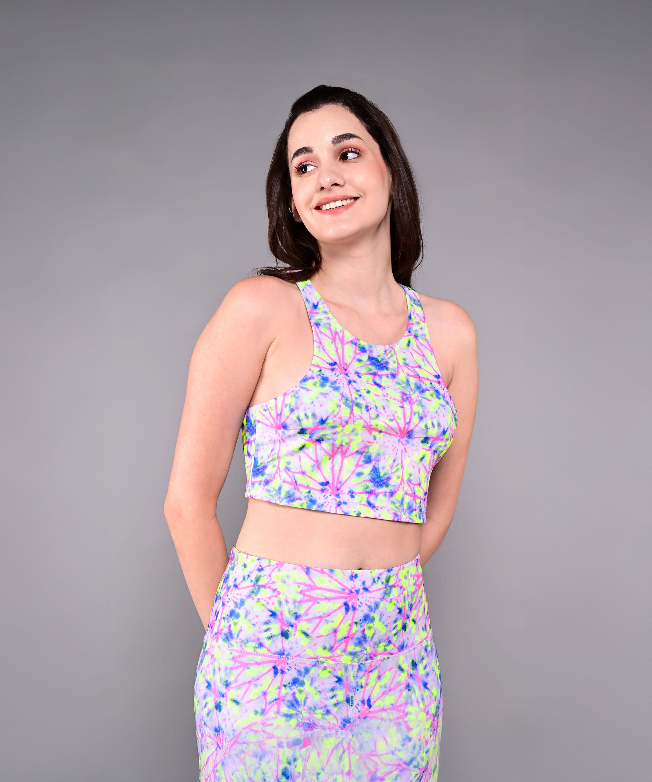 Woman in a Multicolor Neon Crop Top and skirt, smiling with her hands behind her back against a gray background. The outfit is made from skin-friendly cotton lycra for all-day comfort.