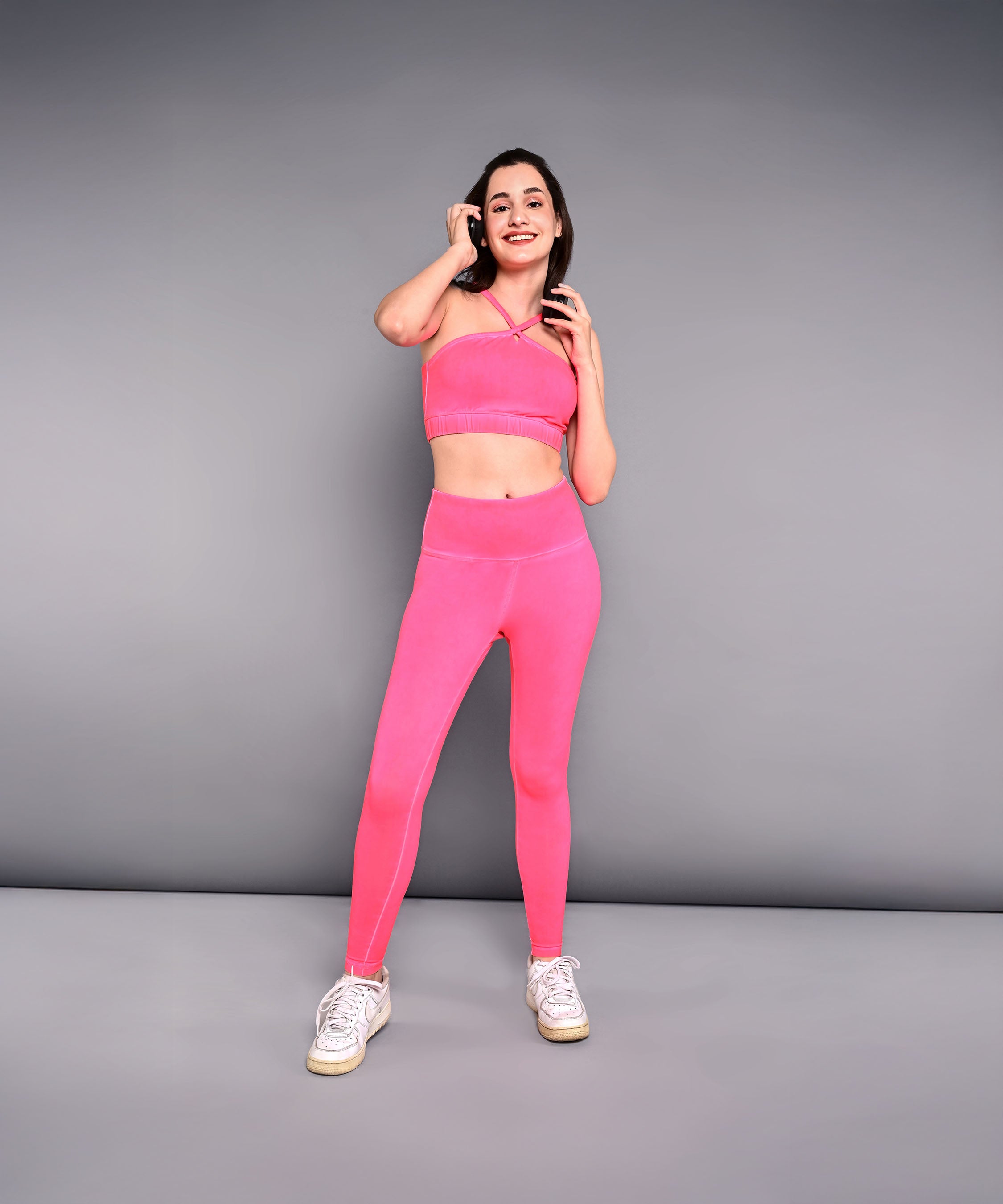 A smiling woman wears the Neon Pink Sports Bra, made from skin-friendly cotton lycra, and poses against a gray wall while holding her hair.